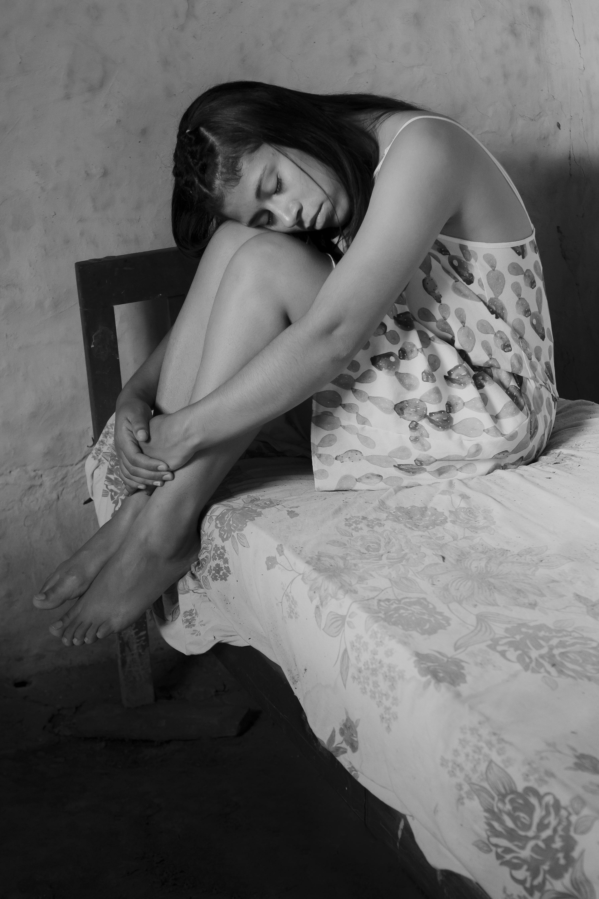 Woman in a patterned dress sitting curled up on a bed, head resting on her knees in a dim room.