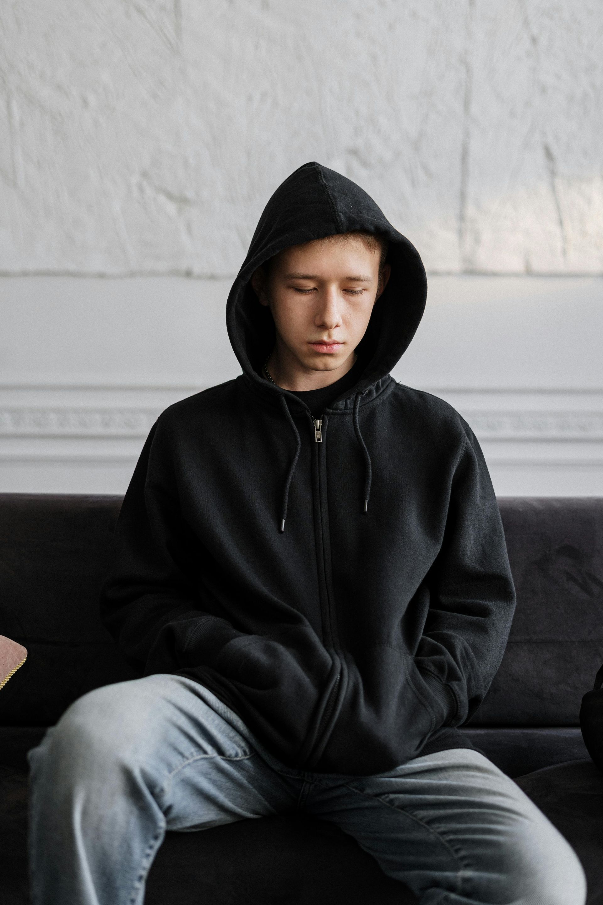 A young man in a black hoodie is sitting on a couch with his eyes closed.