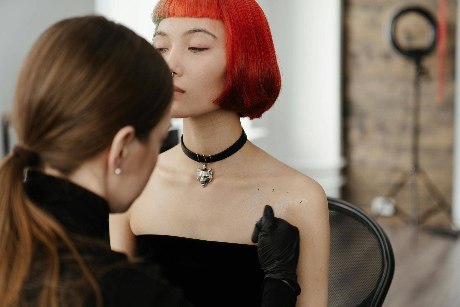 Woman with red hair and choker begins to get a tattoo by a person wearing black gloves in an indoor setting.
