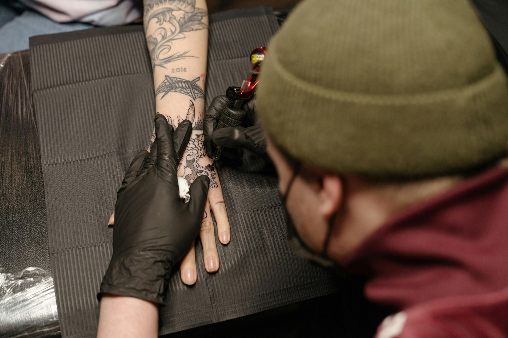 Tattoo artist in black gloves working on a hand and forearm in a dark setting.
