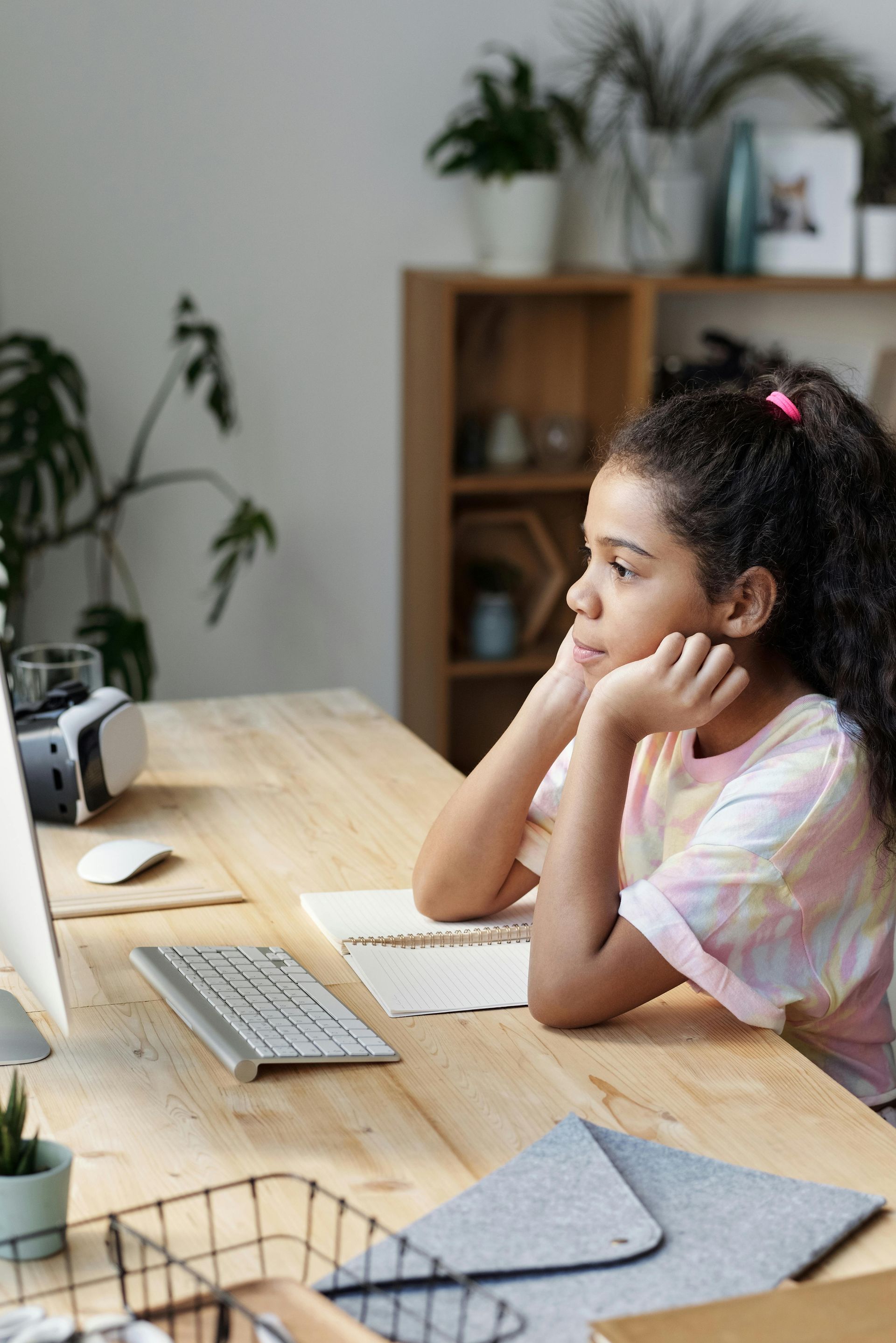 Girl looks at computer screen with hands on her chin, at wooden desk.