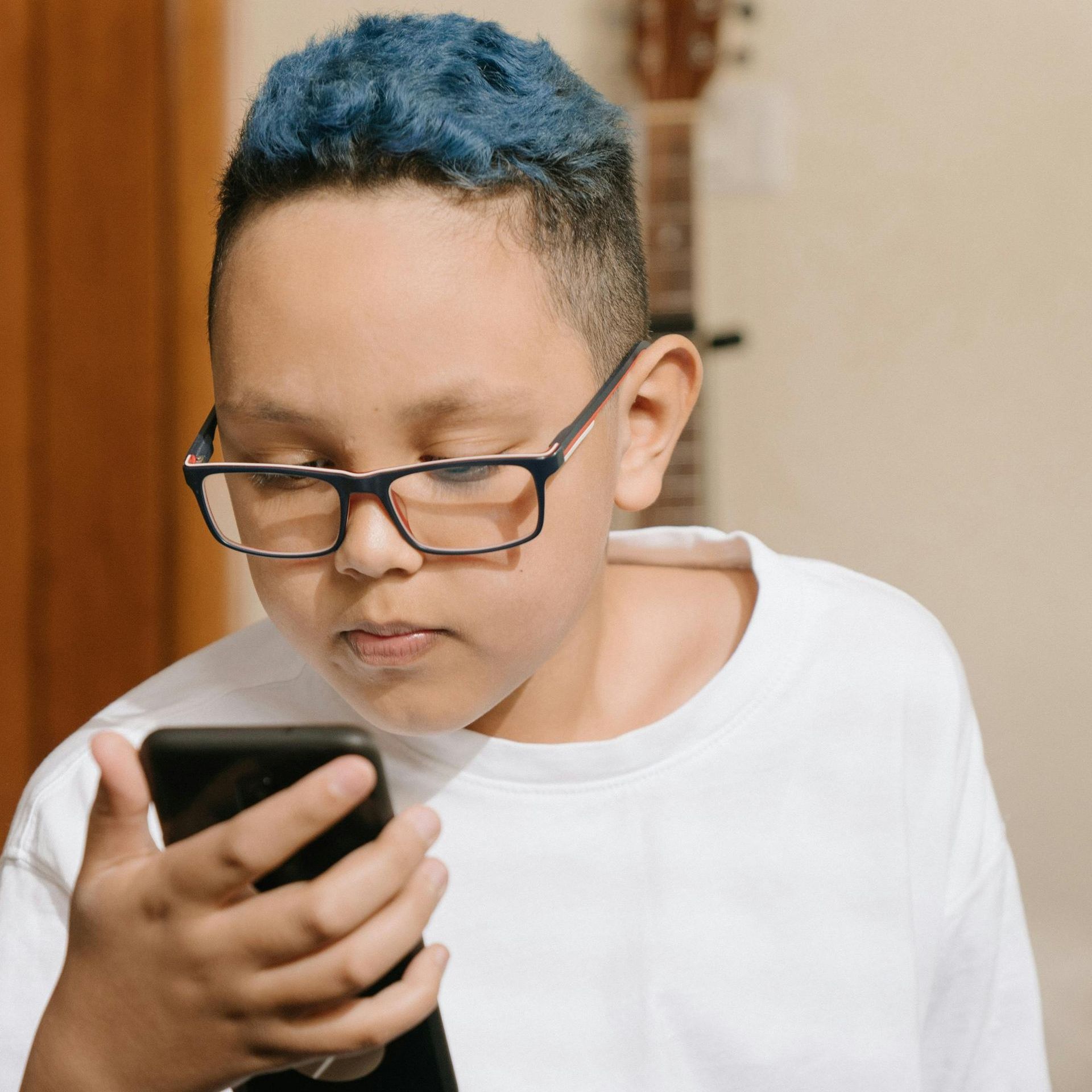 A boy with blue hair and glasses is looking at a cell phone