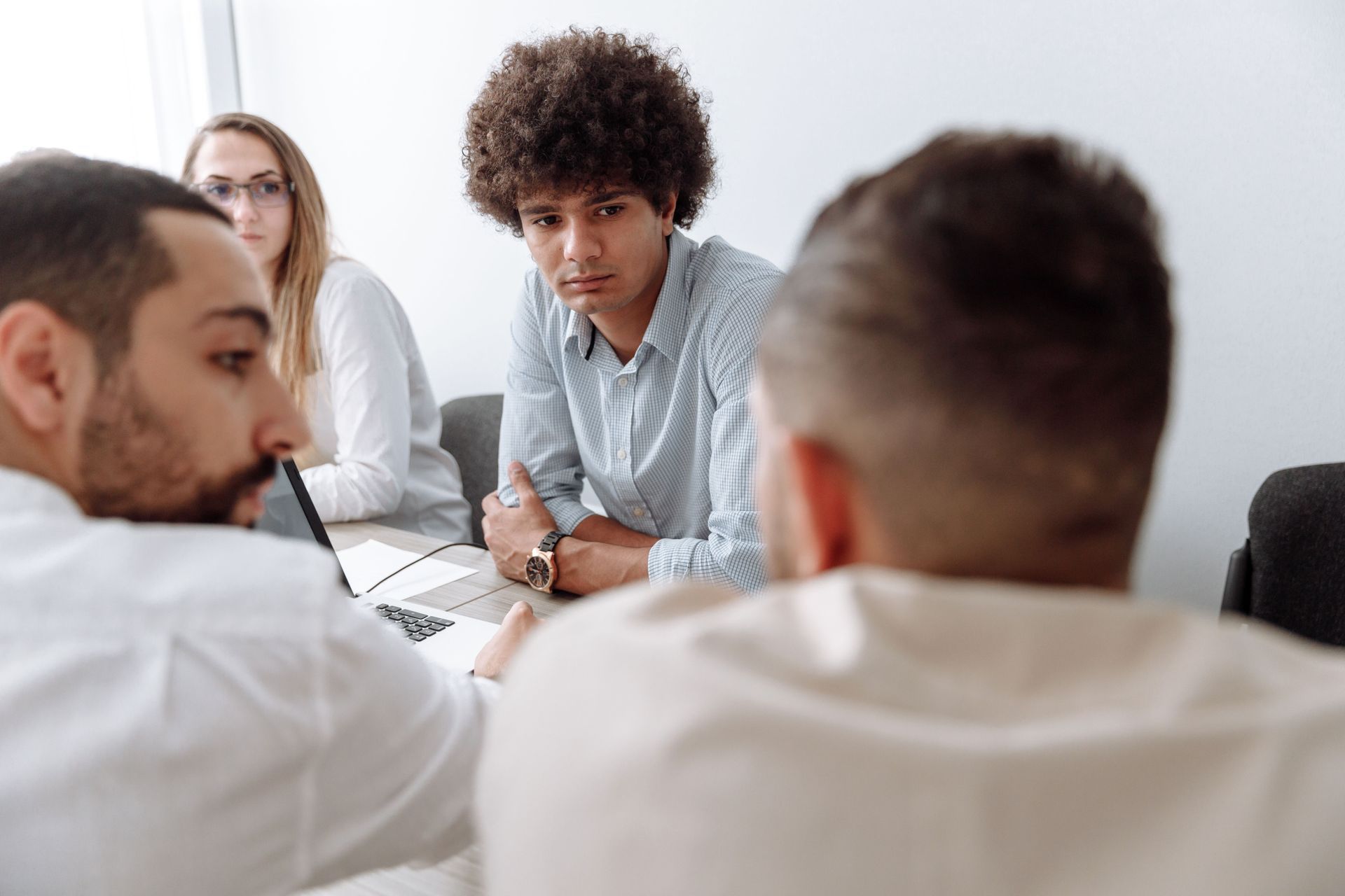 People in a meeting around a table, listening to a speaker in a bright room.