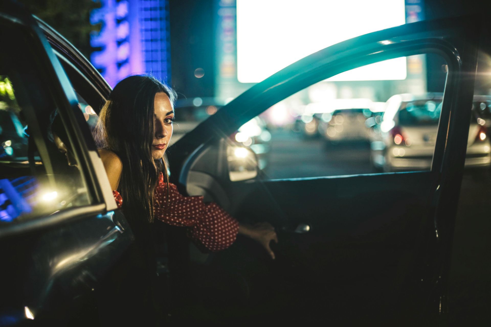 Woman in red polka-dot top leans out of a car door at night, looking toward the camera. Cars in the background.