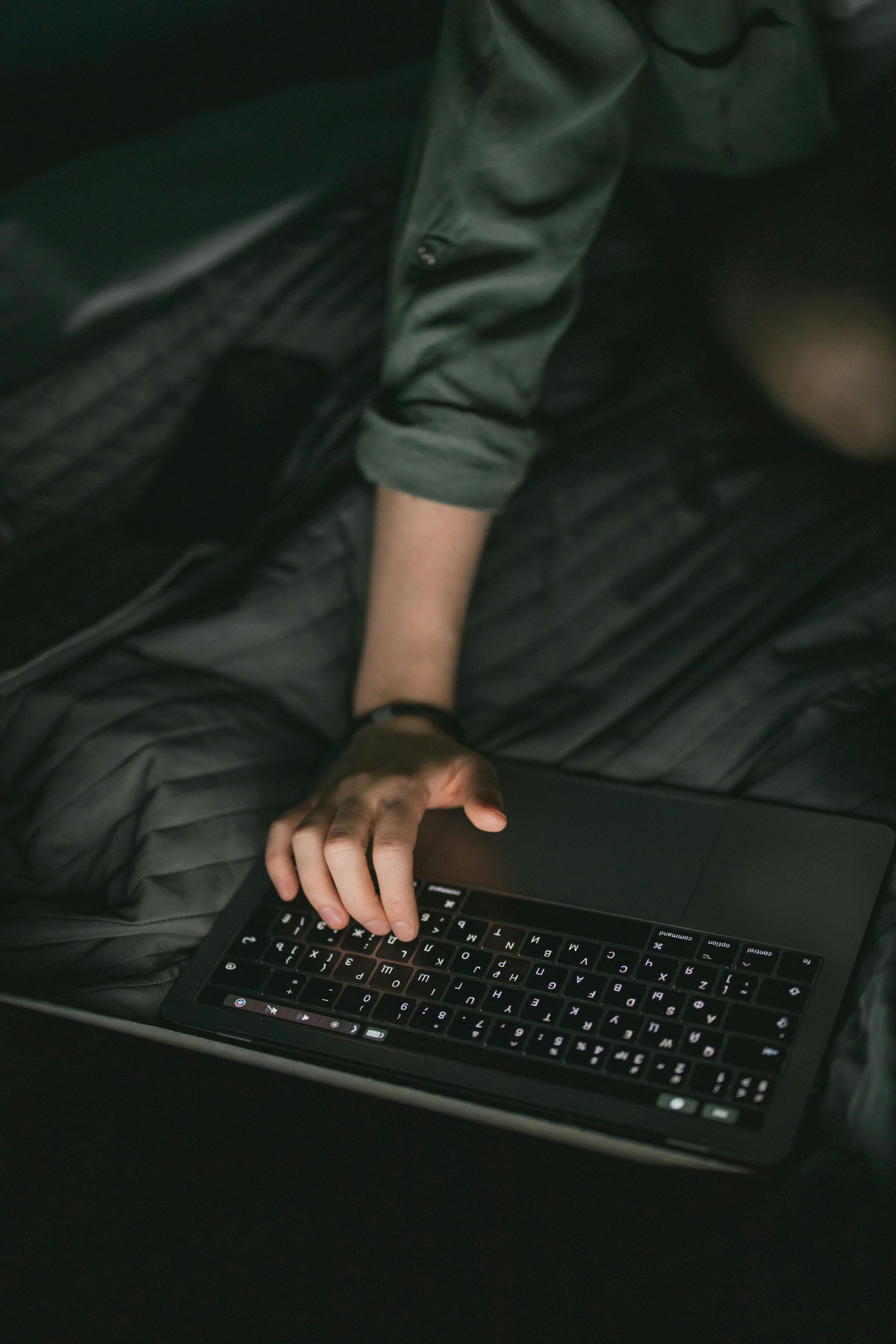A person wearing a gray long-sleeved shirt typing on a laptop computer while sitting on a dark, quilted surface.