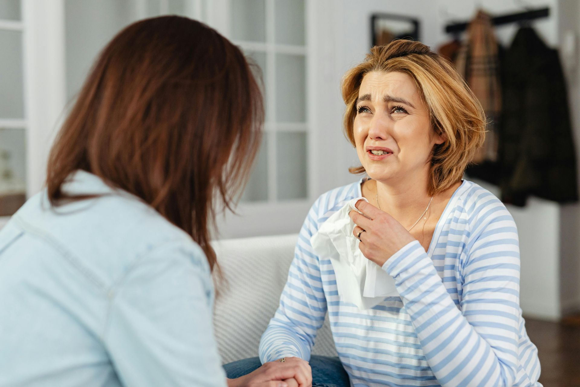 Two women sitting indoors, holding hands, one looking upset and crying during a conversation
