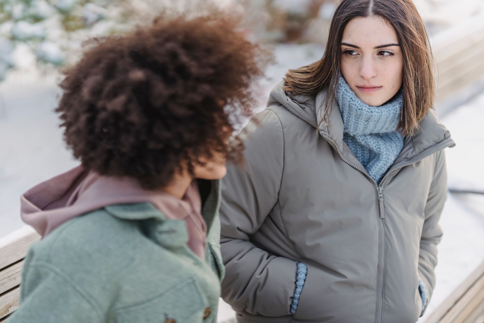 Two people talking on a snowy bench, one in a gray coat and blue scarf, the other in a green jacket.