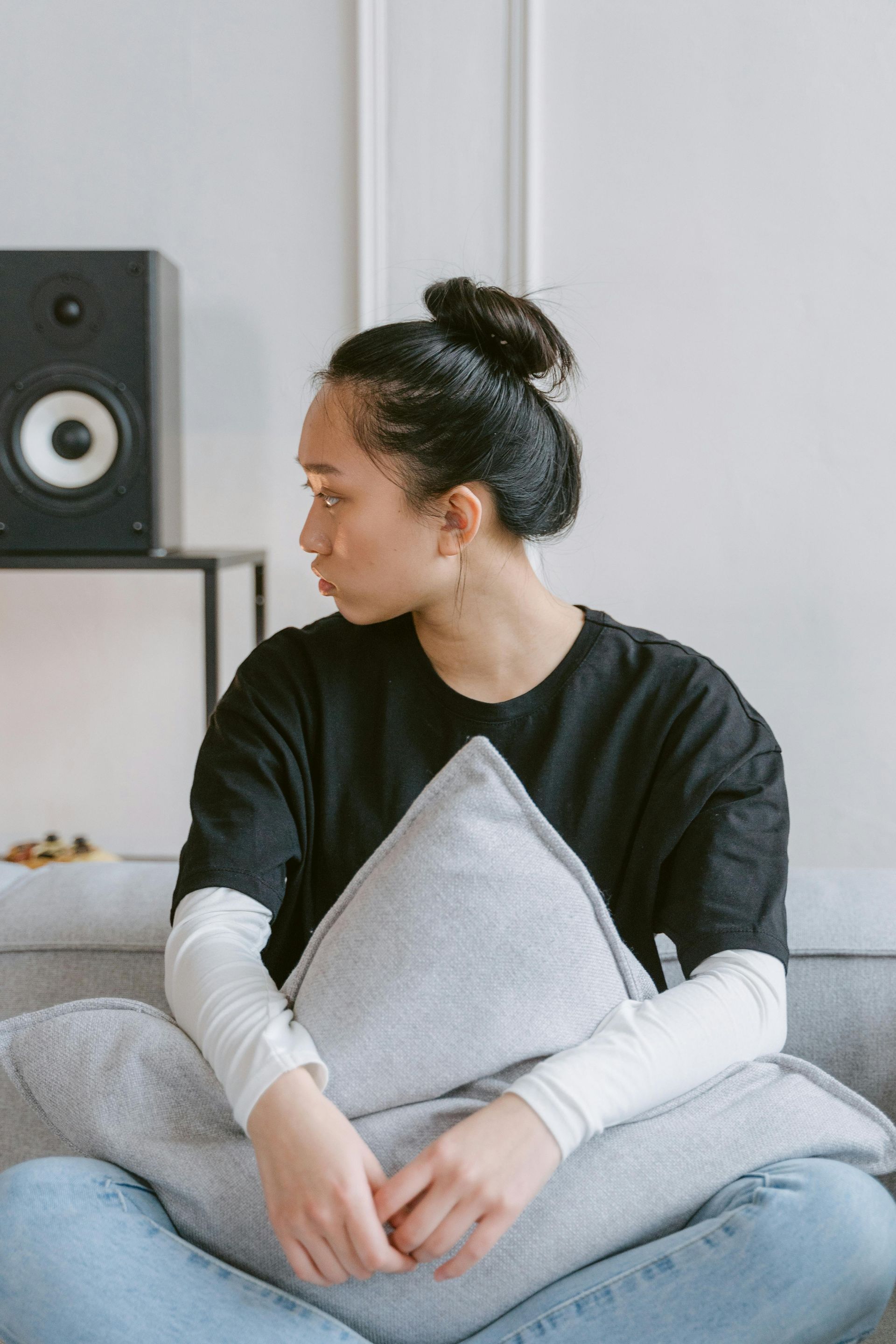 Person sitting cross-legged on a couch holding a gray pillow, looking to the side in a bright room.