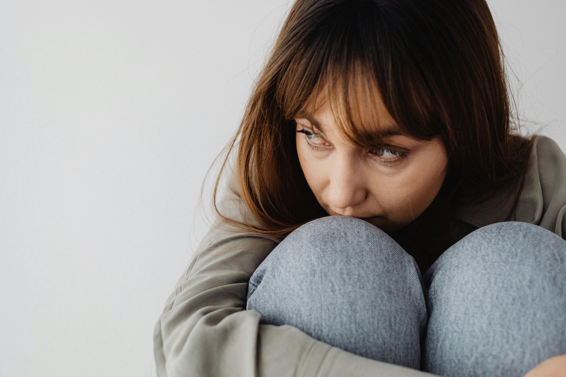 Woman sitting curled up against a white wall, wearing a gray sweater and jeans, looking down sadly