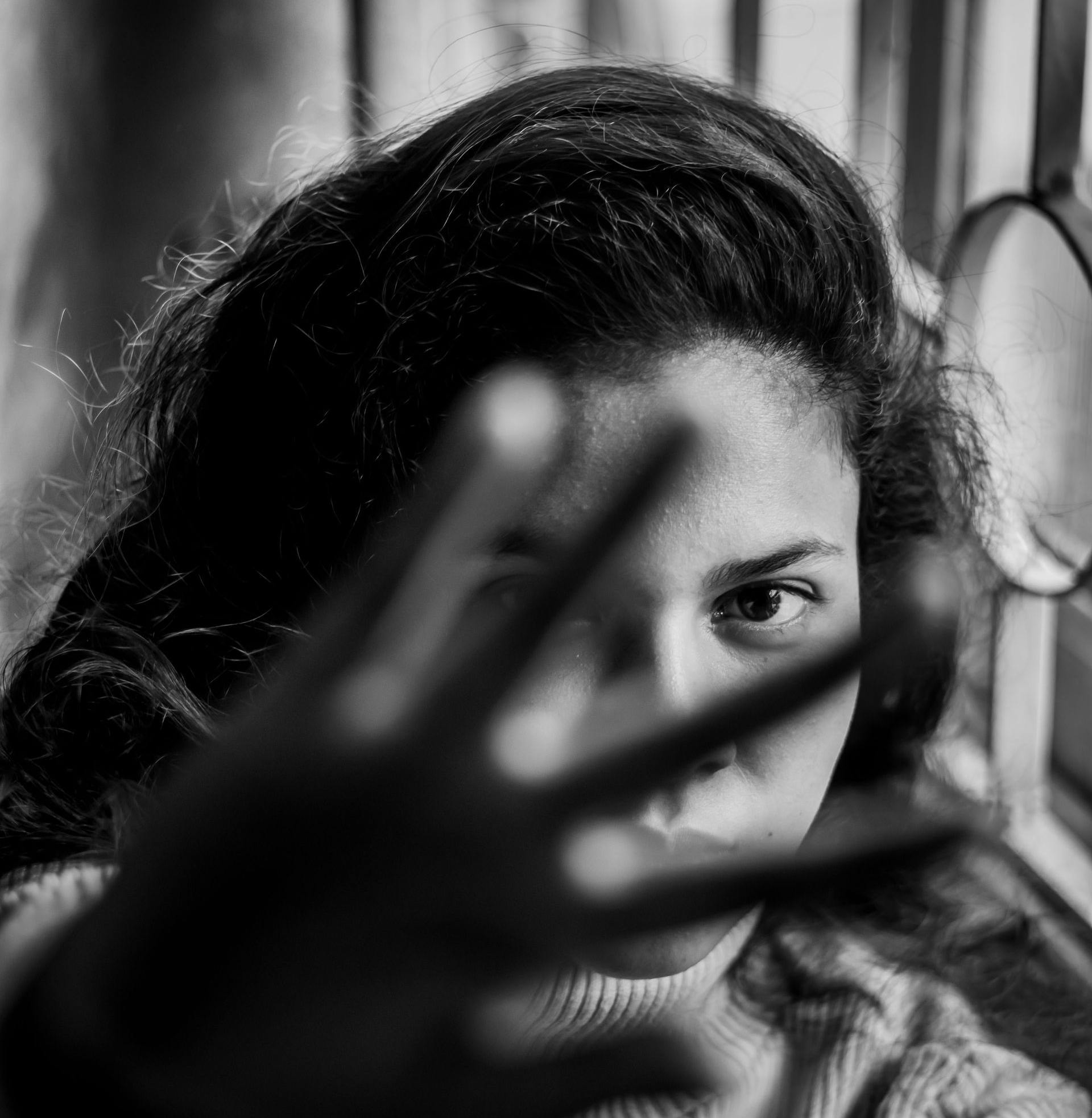 Black-and-white portrait of a person peering through an outstretched hand near a window