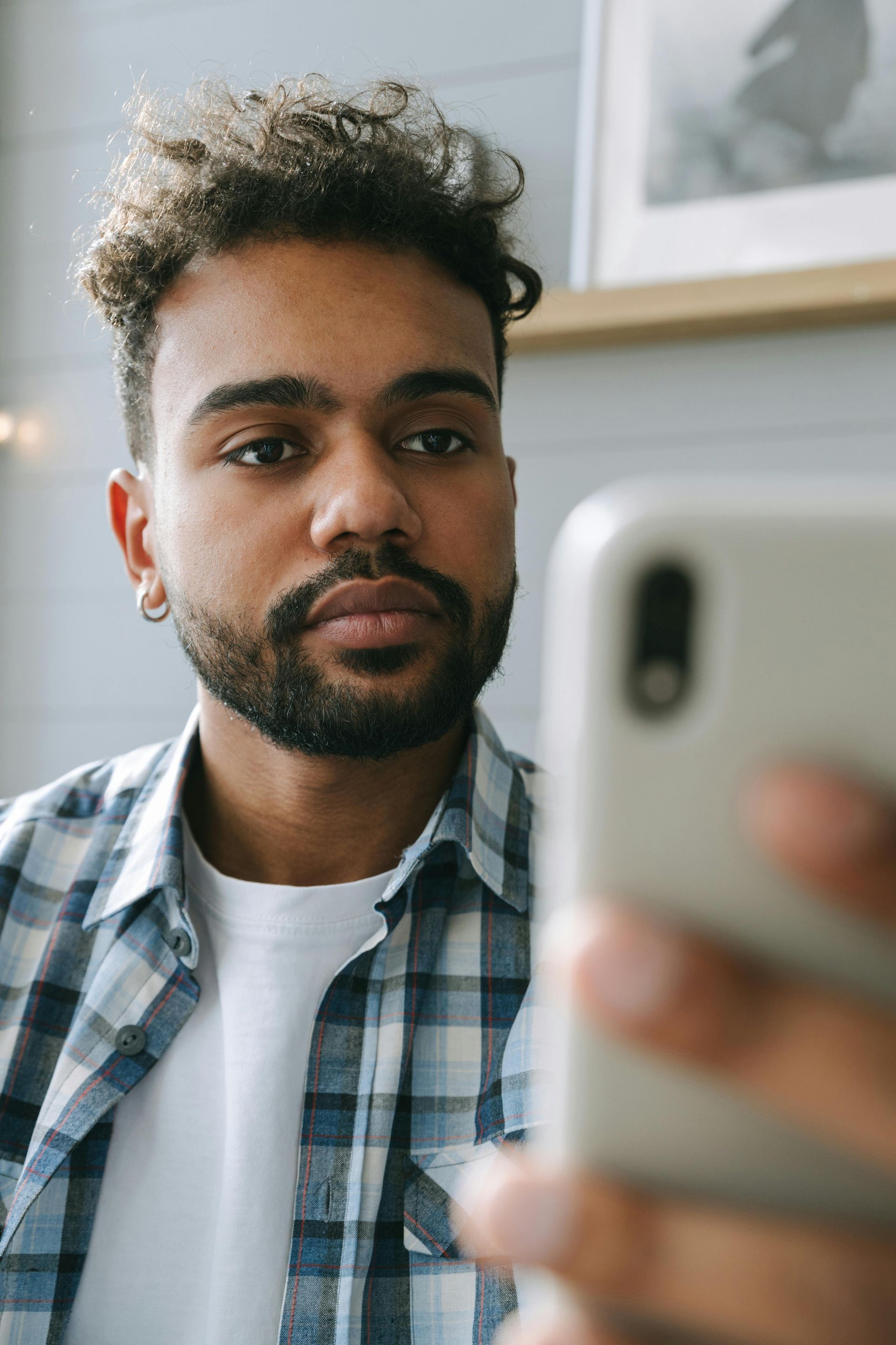 A man with curly hair and a beard looks at his phone. He wears a plaid shirt and a silver earring.