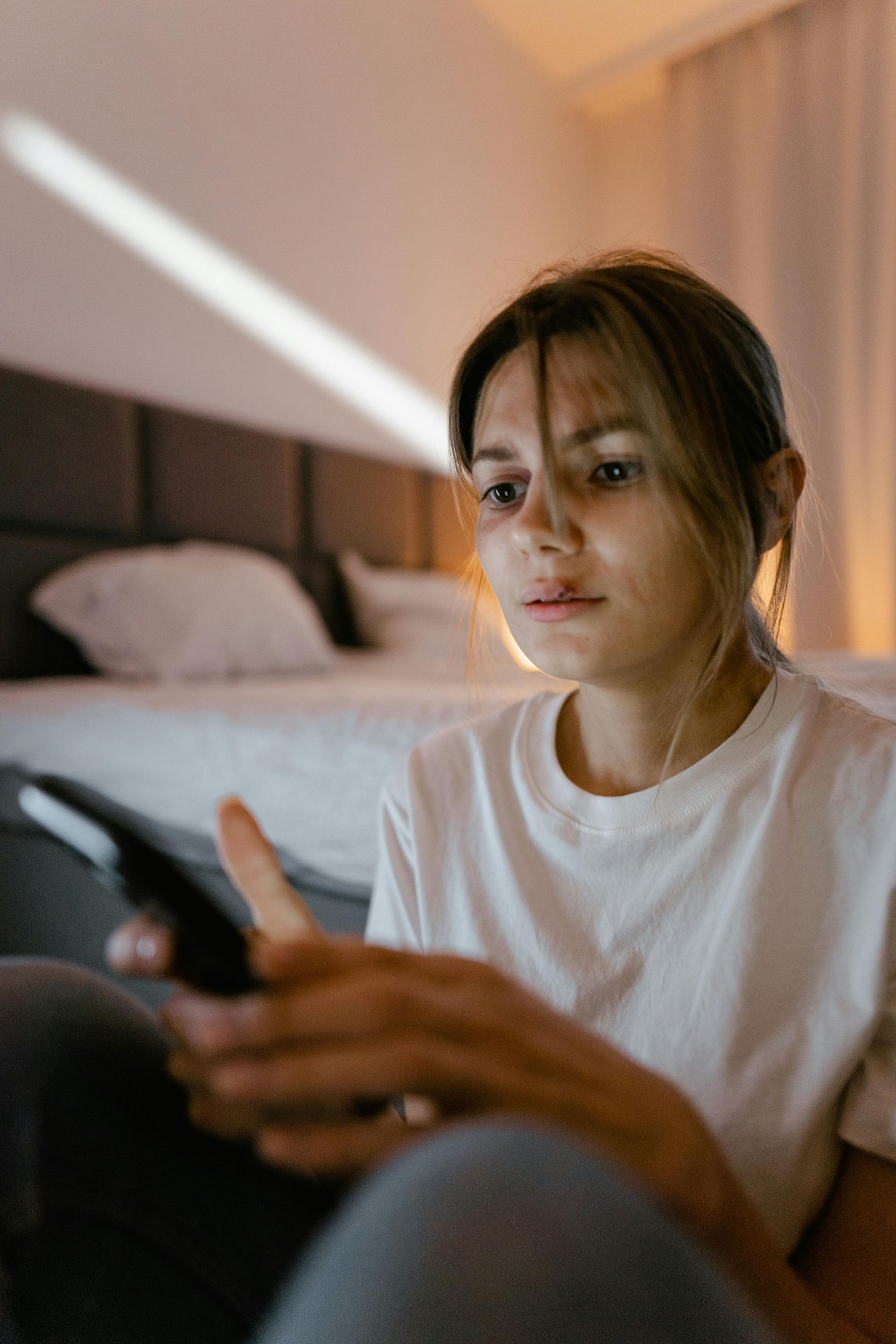 Woman in white shirt using a phone, sitting on floor in front of a bed.