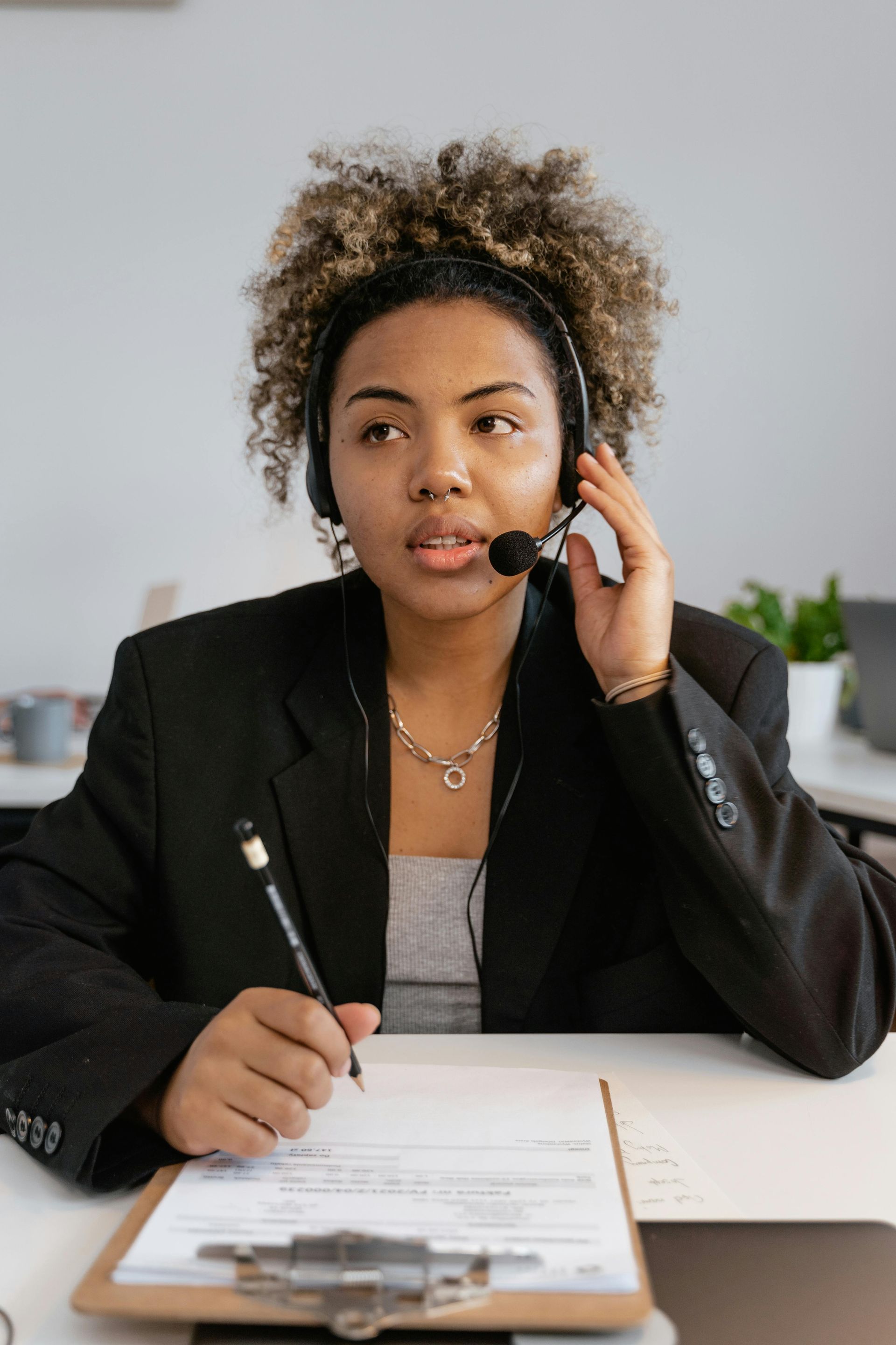 Woman with headset, writing on clipboard, in office setting.