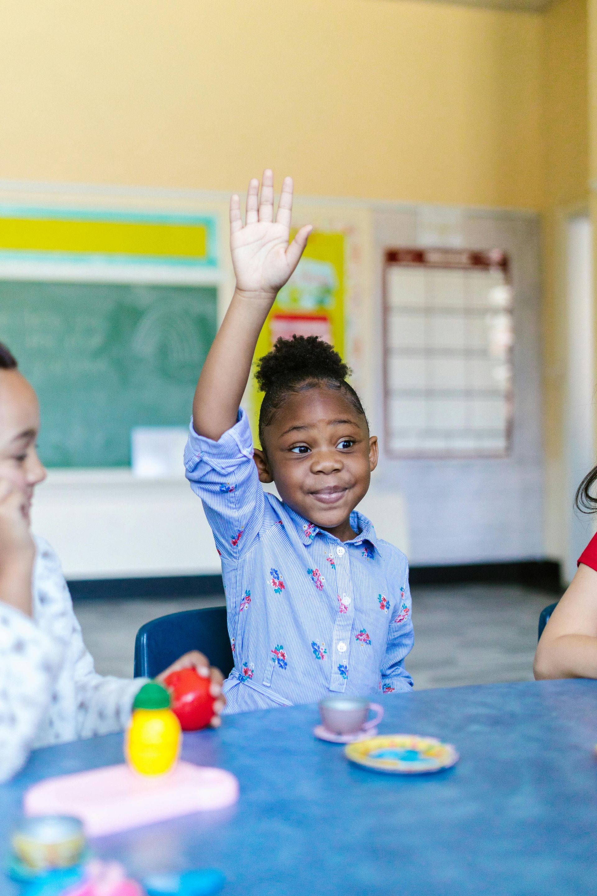 Young Black girl raises hand in classroom; smiling, blue shirt, toys on table.