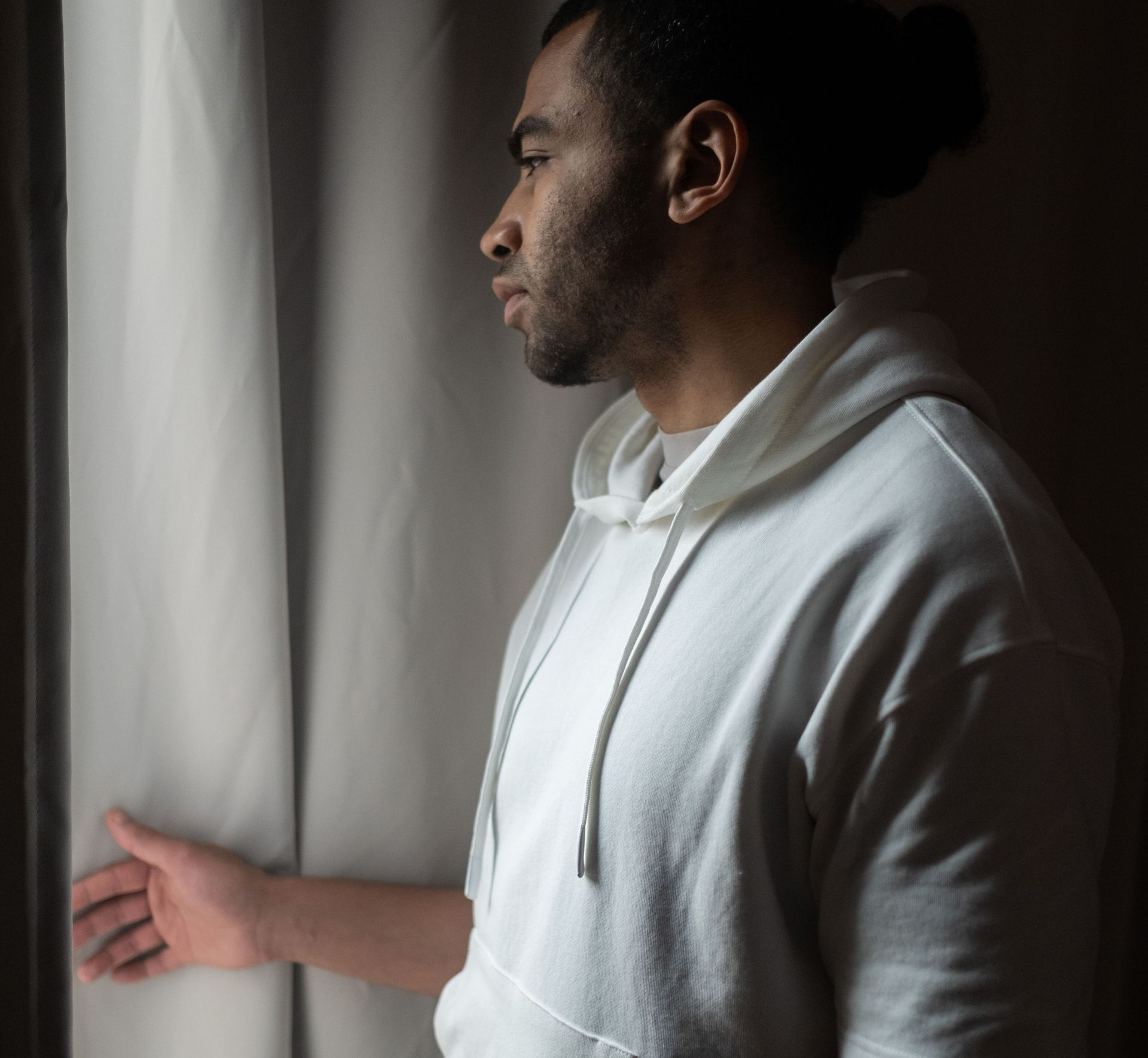 Person in a white hoodie standing by a window, looking out with a thoughtful expression.