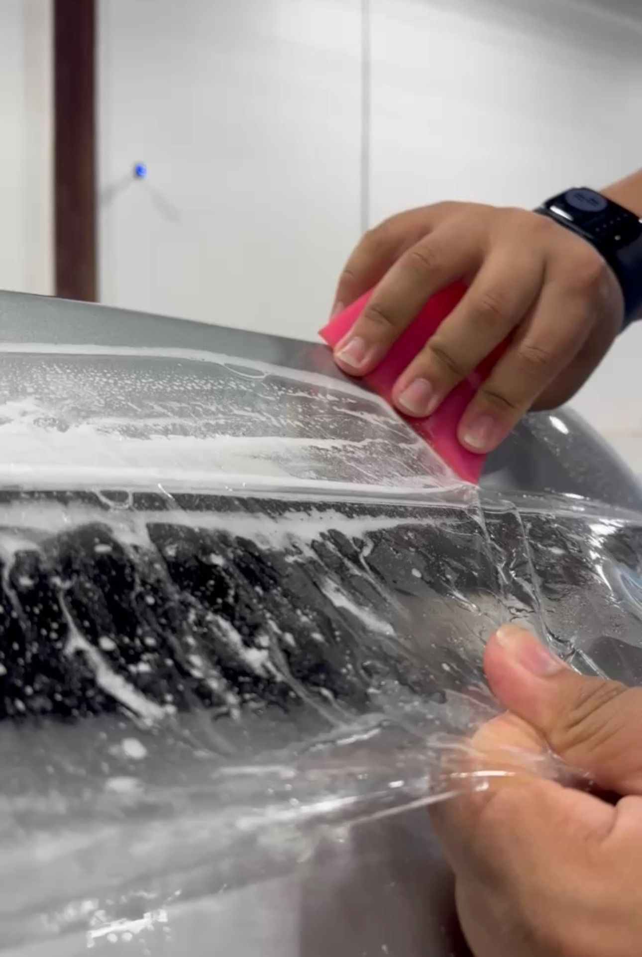 Person using a sponge to smooth clear film on a car's surface. Soapy water visible.