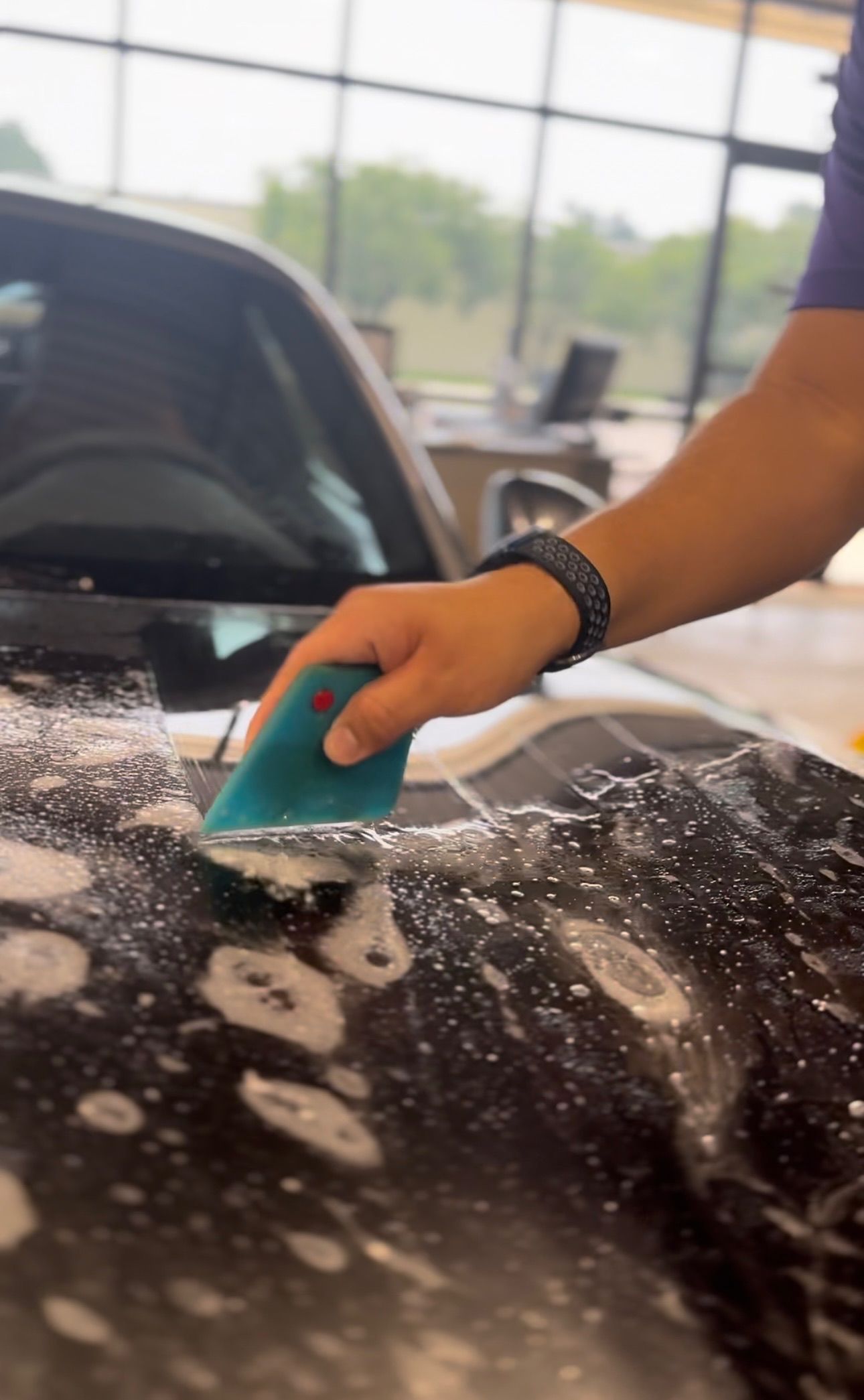Person using a squeegee to apply film to a car's hood, which is covered in soapy water.