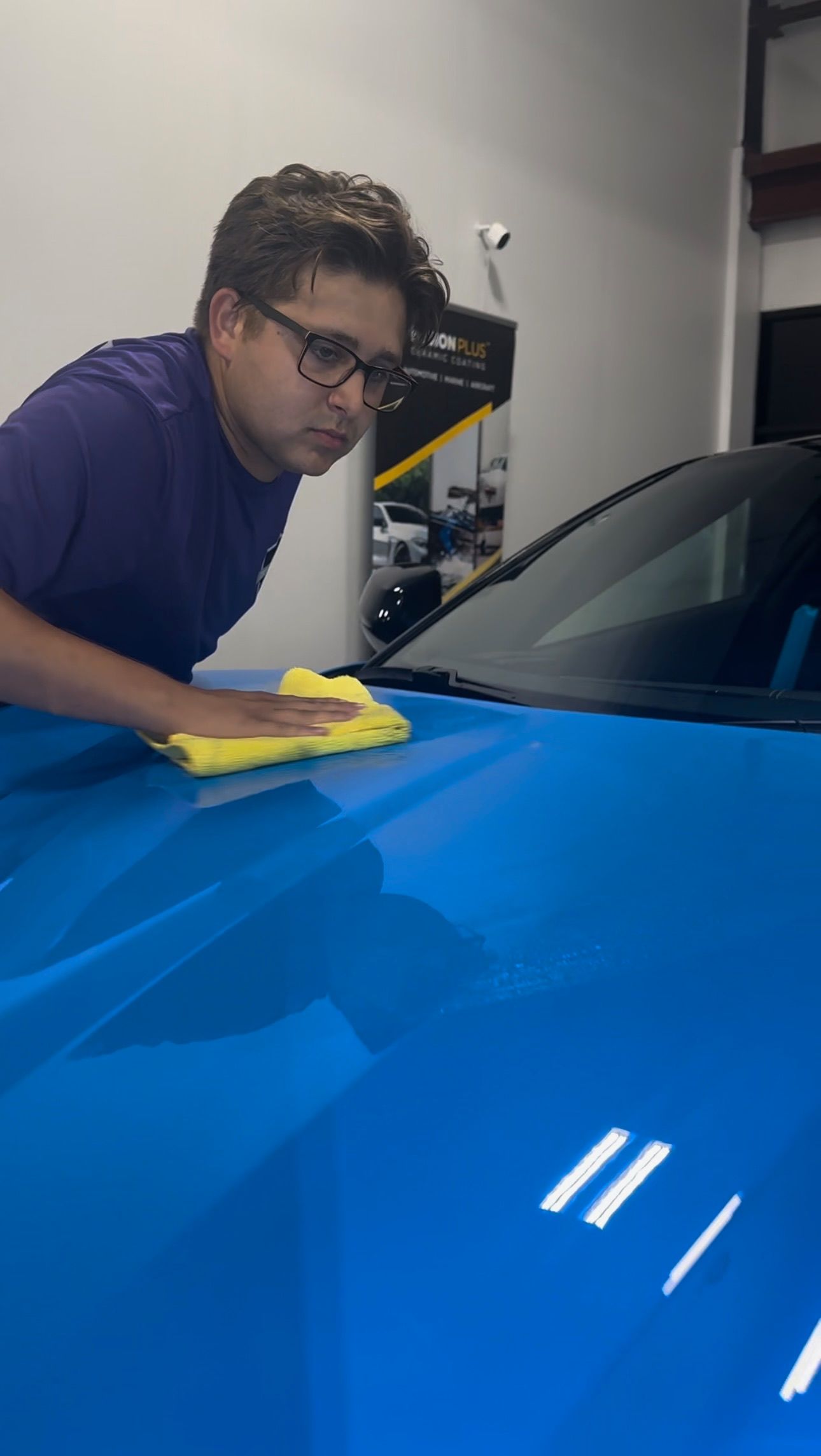 Person wiping a blue car's hood with a yellow cloth. In a garage-like setting.