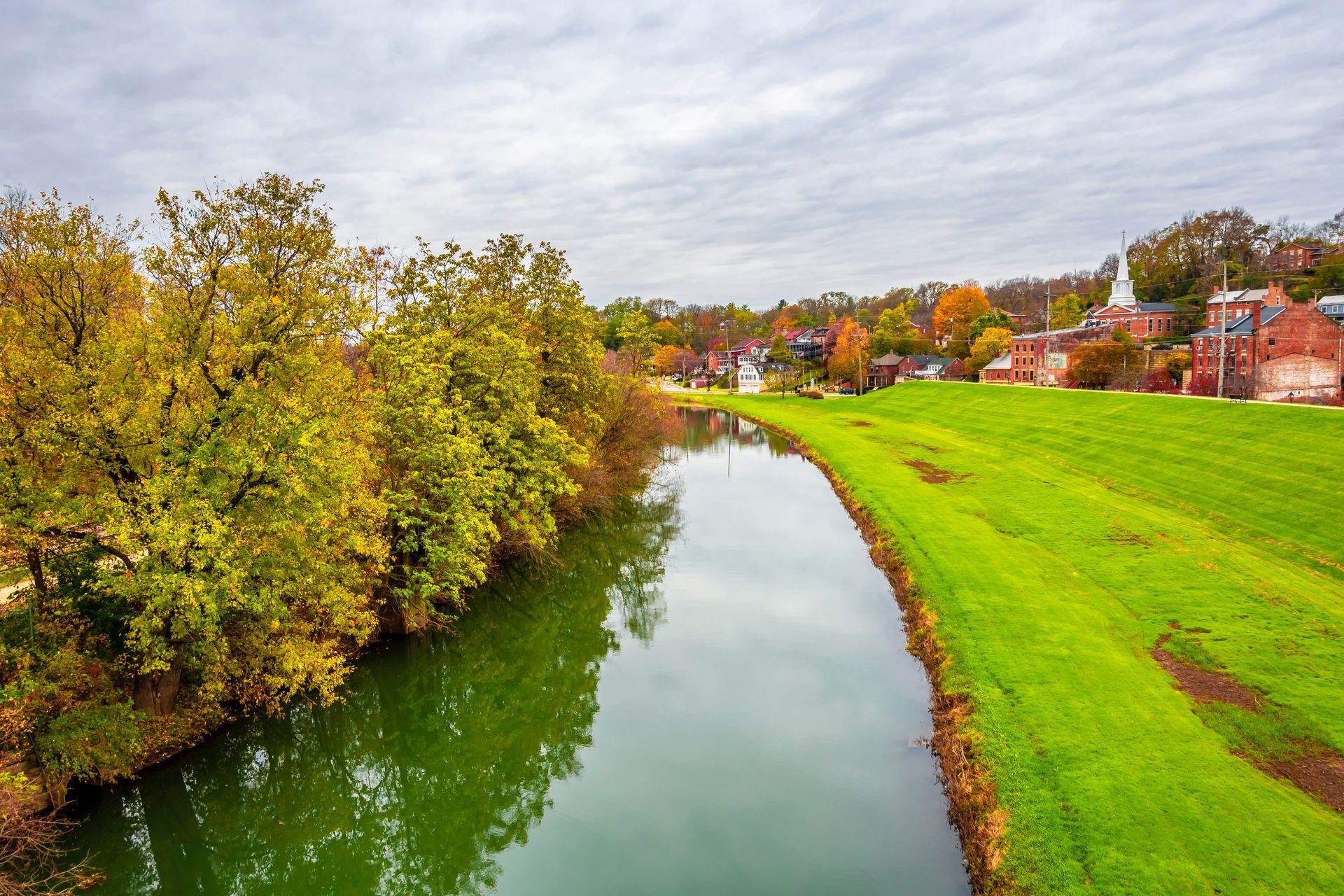 A river flows through a grassy embankment lined with trees displaying autumn colors, with buildings in the background.