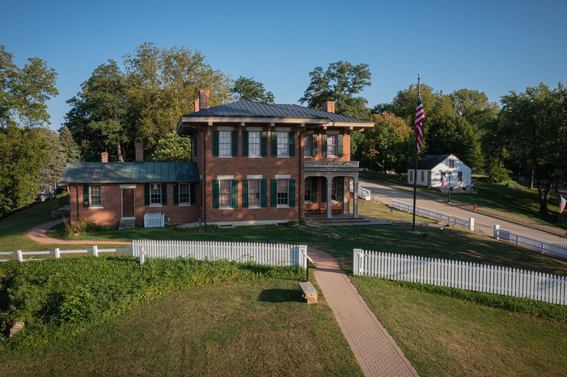Two-story brick house with green shutters, white picket fence, flagpole, and small white building on a hill.