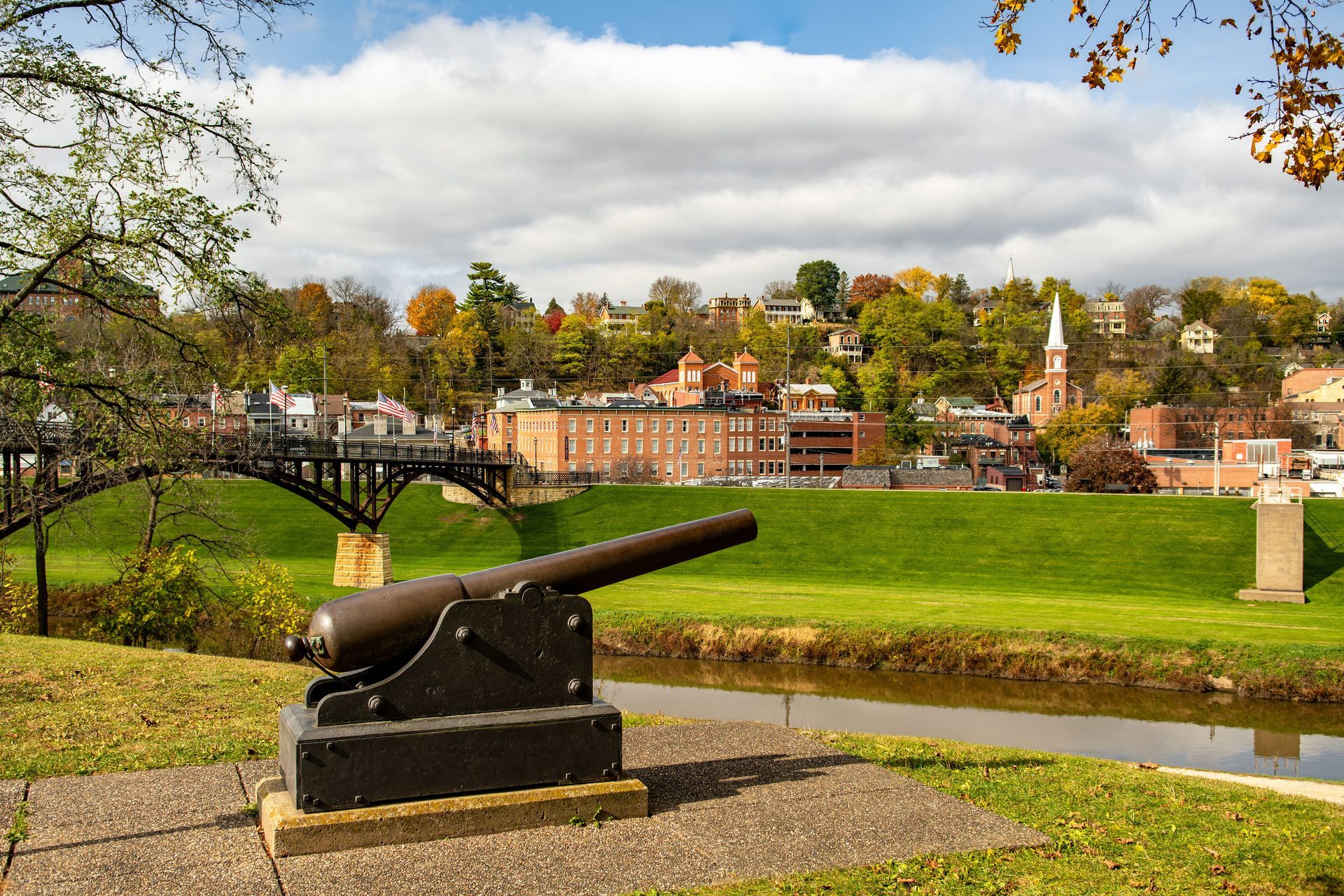 Cannon overlooking a town, bridge, and river, set against a backdrop of trees and a cloudy sky.
