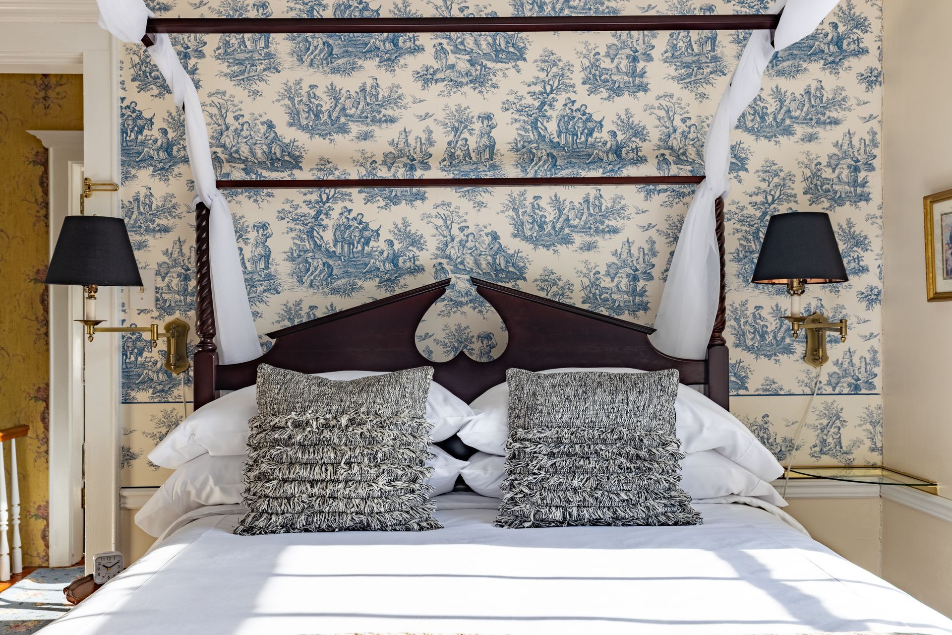 A bed with a wooden frame and canopy, blue and white wallpaper, two decorative pillows, and black lamps.