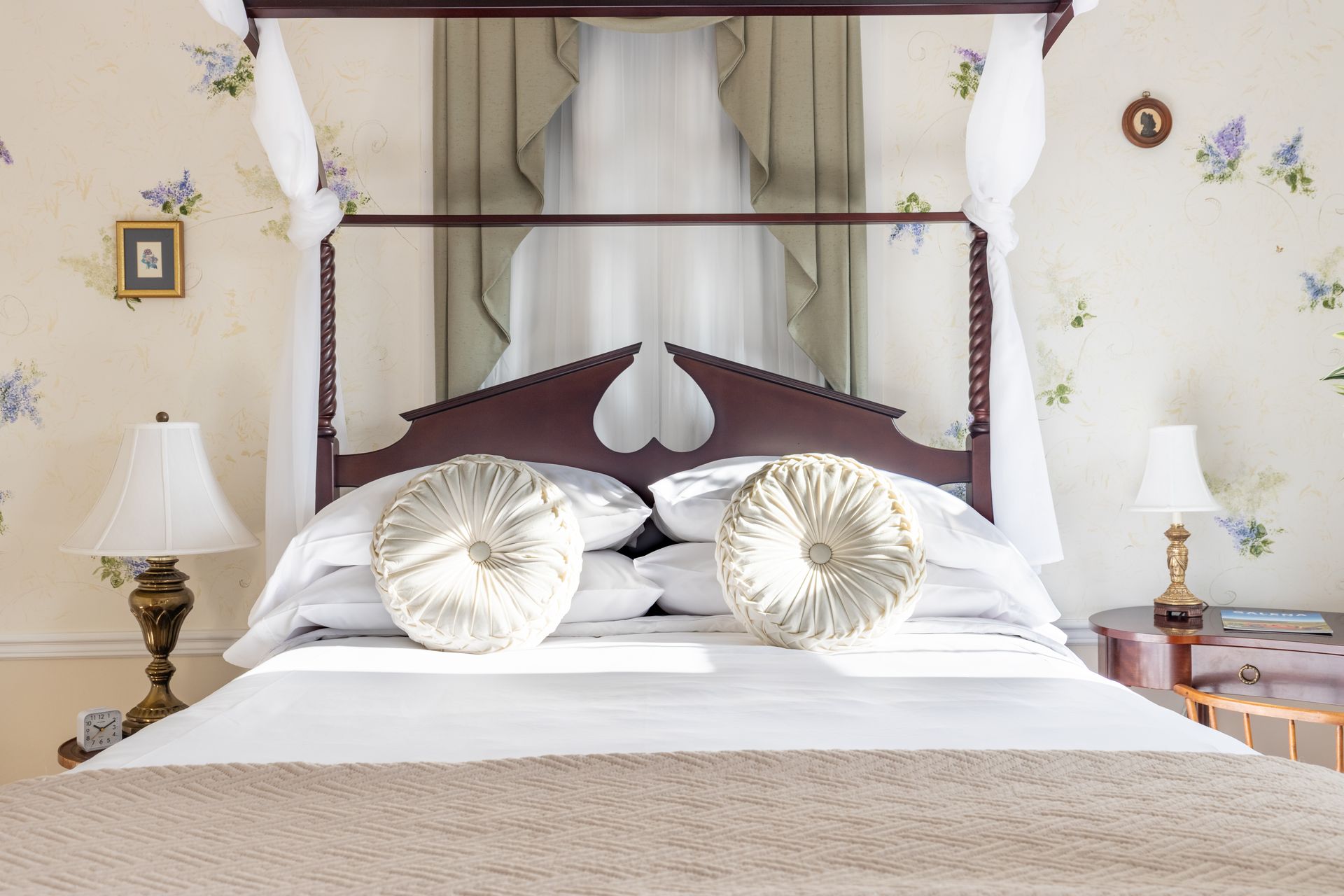 Bedroom with four-poster bed, floral wallpaper, and two round pillows. Two lamps on nightstands on either side.