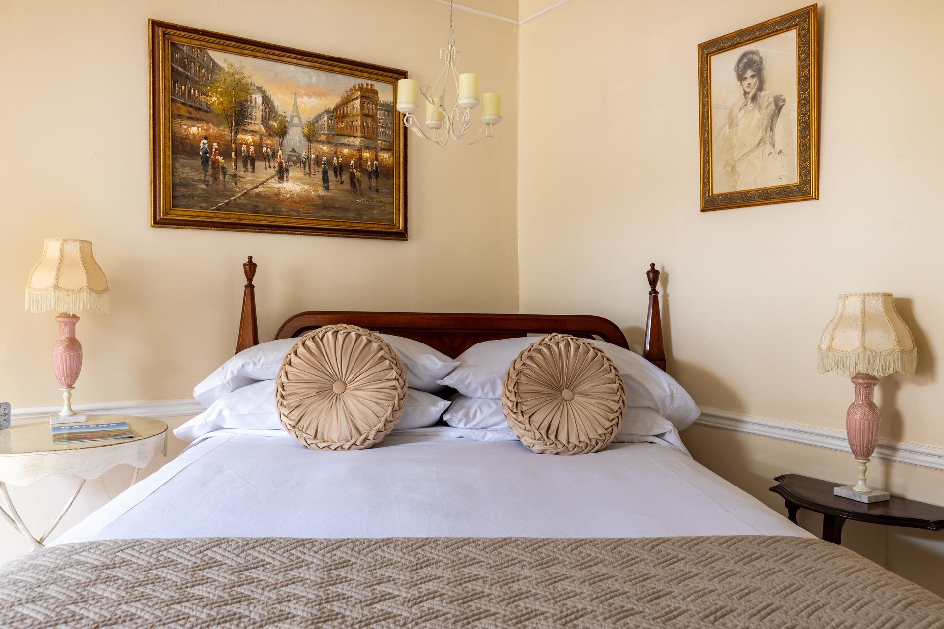 Bedroom with a bed, lamps, and artwork. Beige walls, wood bed frame, two decorative pillows, and a painting above the bed.