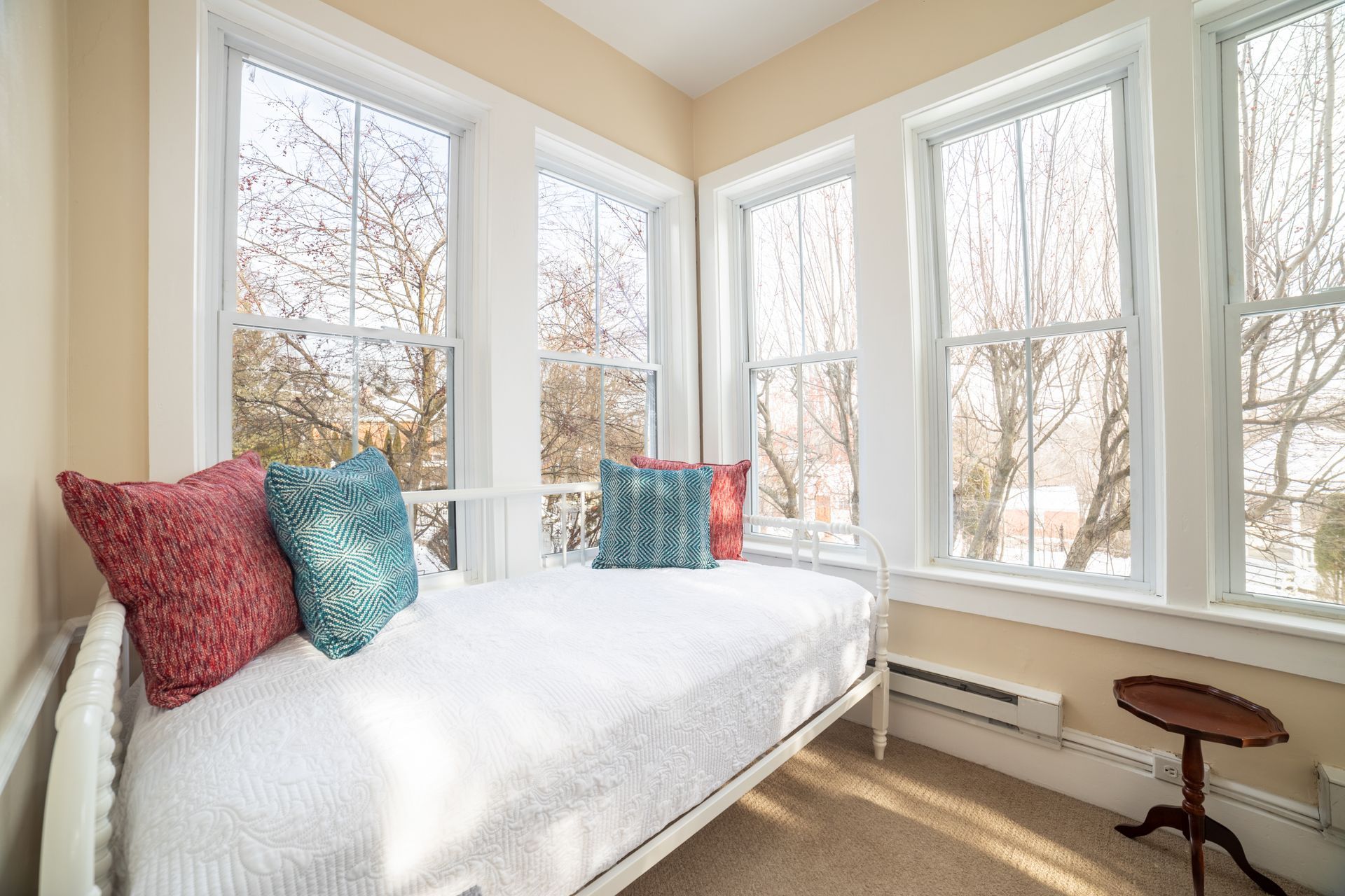 Cozy sunroom with white daybed, three windows, and small table. Red and teal pillows against a bright backdrop.