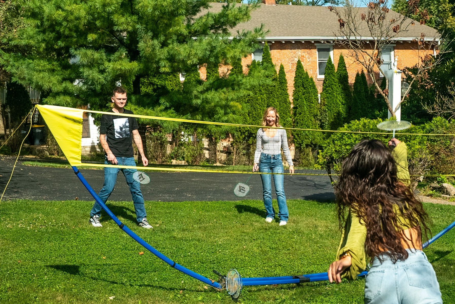 People playing badminton in a yard with a net. The sky is bright and the house is in the background.