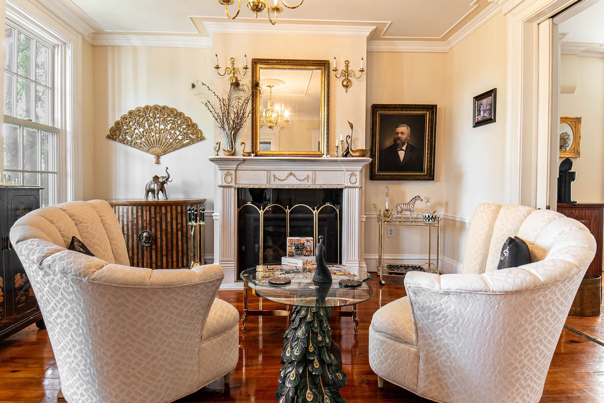 Living room with two white armchairs facing a fireplace, antique furniture, and a mirror.