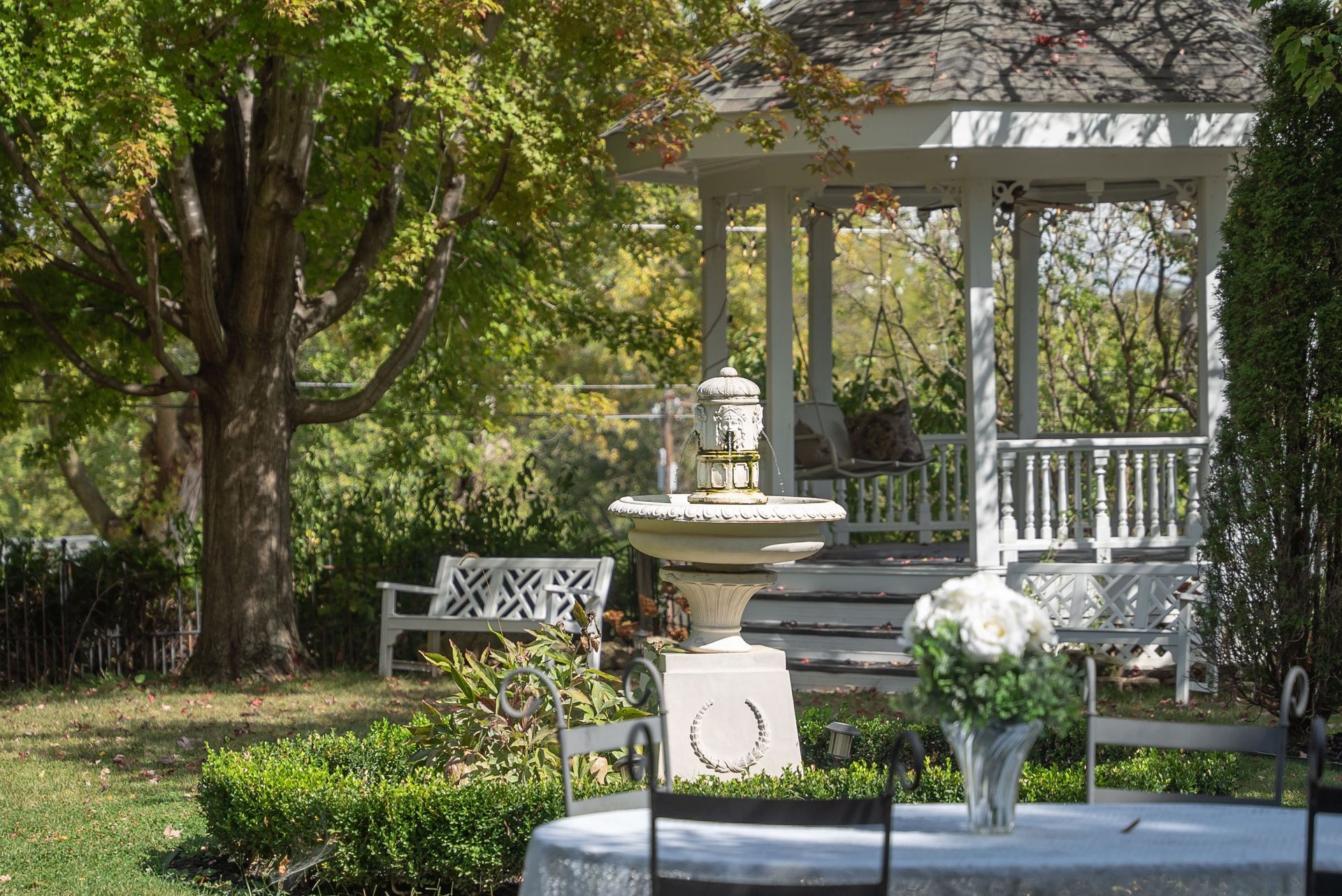 A garden scene with a white gazebo, fountain, and table set for a gathering.