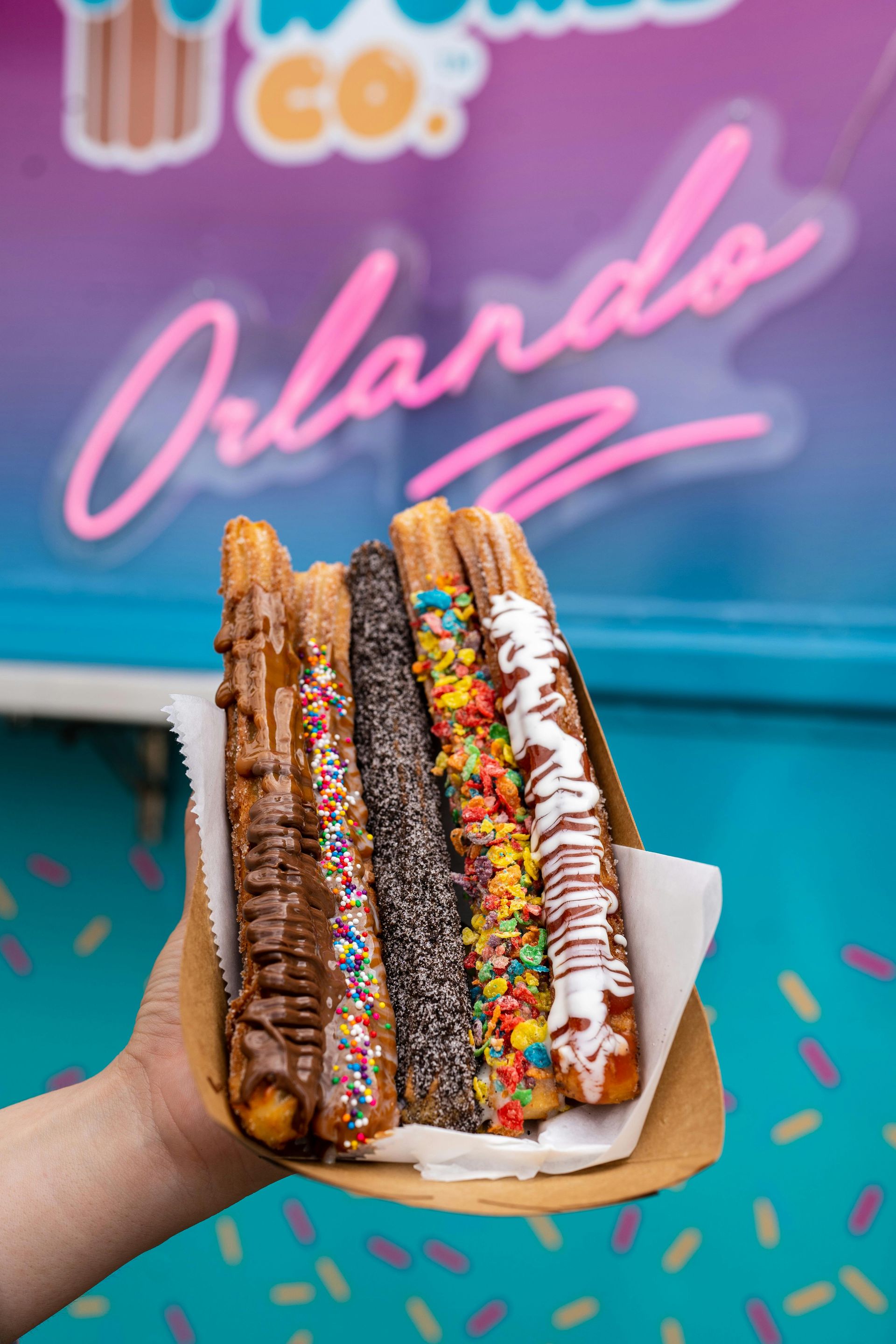 Hand holding a tray of churros with various toppings, in front of a turquoise food truck sign,