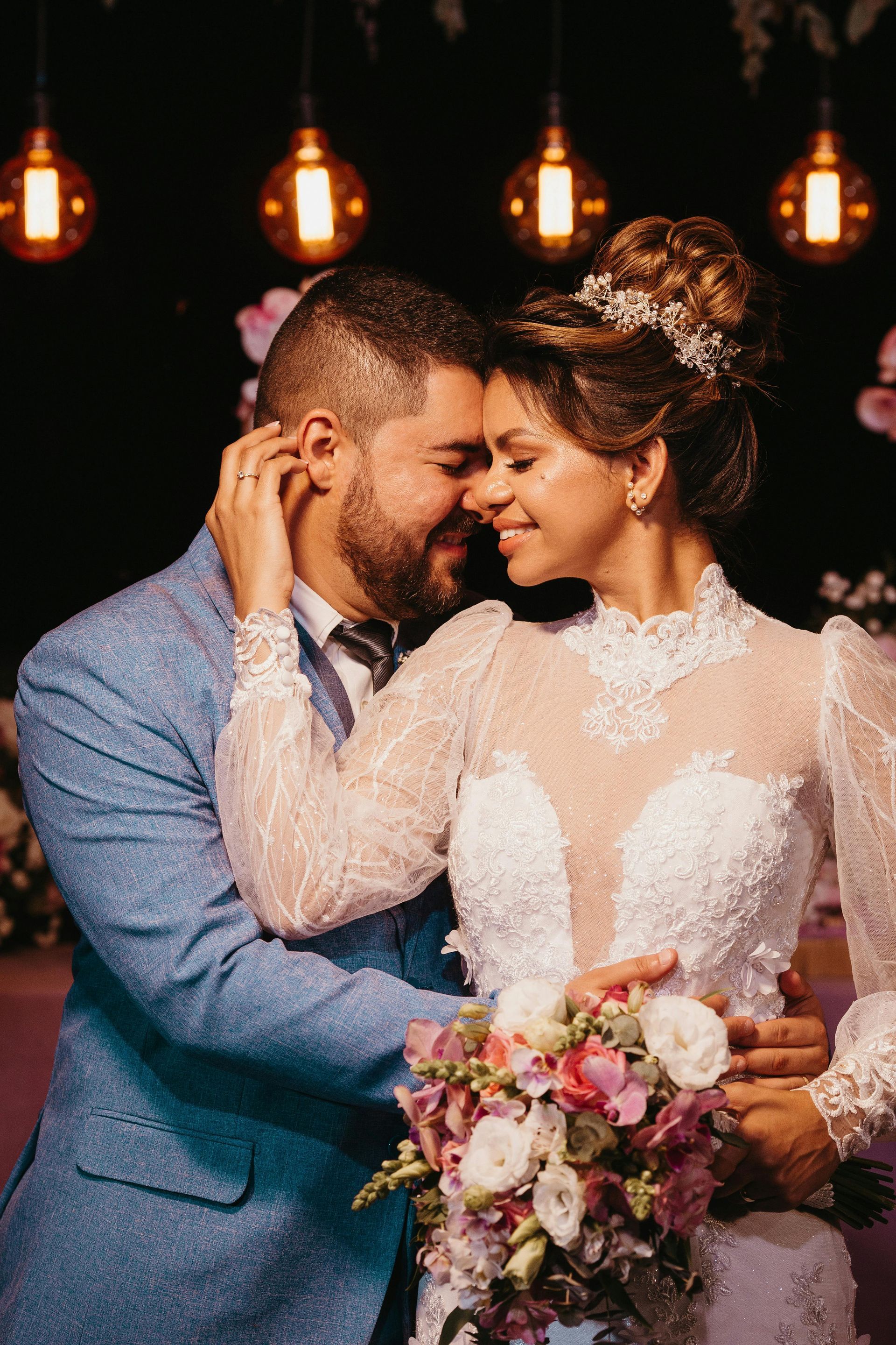 Bride and groom embrace, smiling, at a wedding reception. She wears a white gown with a bouquet. Soft, warm lighting.