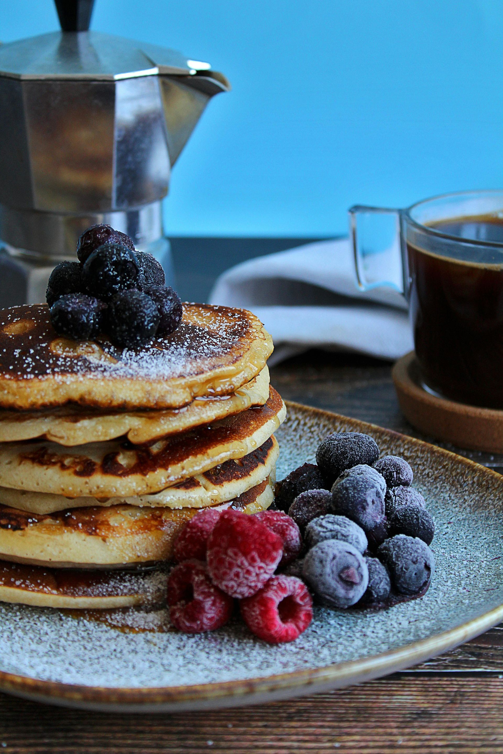 Stack of pancakes topped with berries, dusted with powdered sugar, served with coffee and coffee pot.