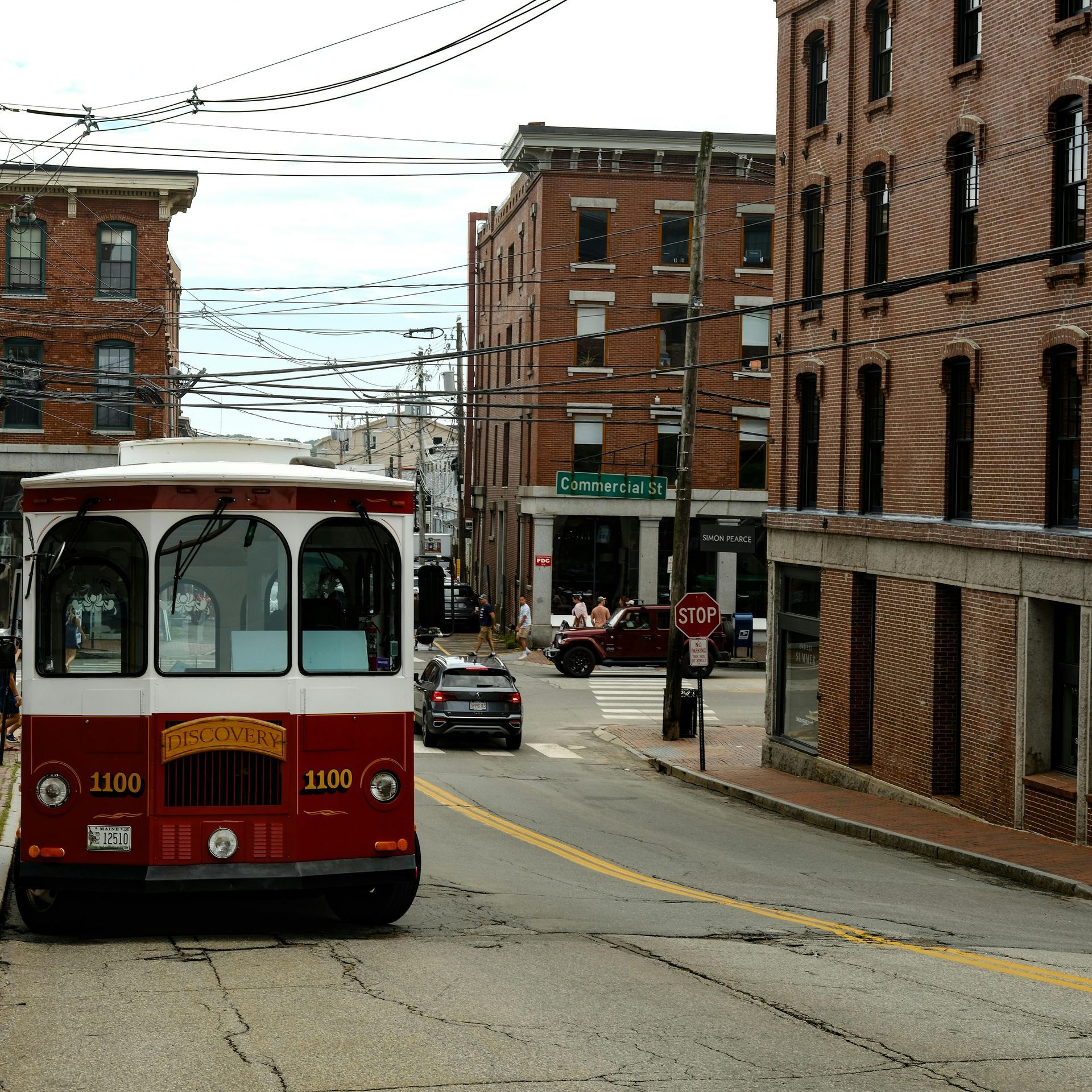 A red and white trolley on a city street, with brick buildings, power lines, and a crosswalk.