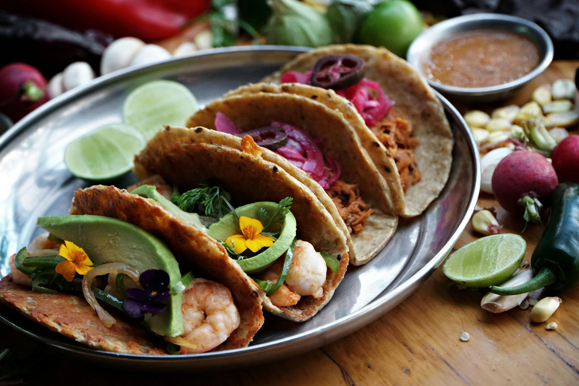Tacos on a silver platter with various toppings: shrimp, avocado, pickled onions, salsa, and limes.