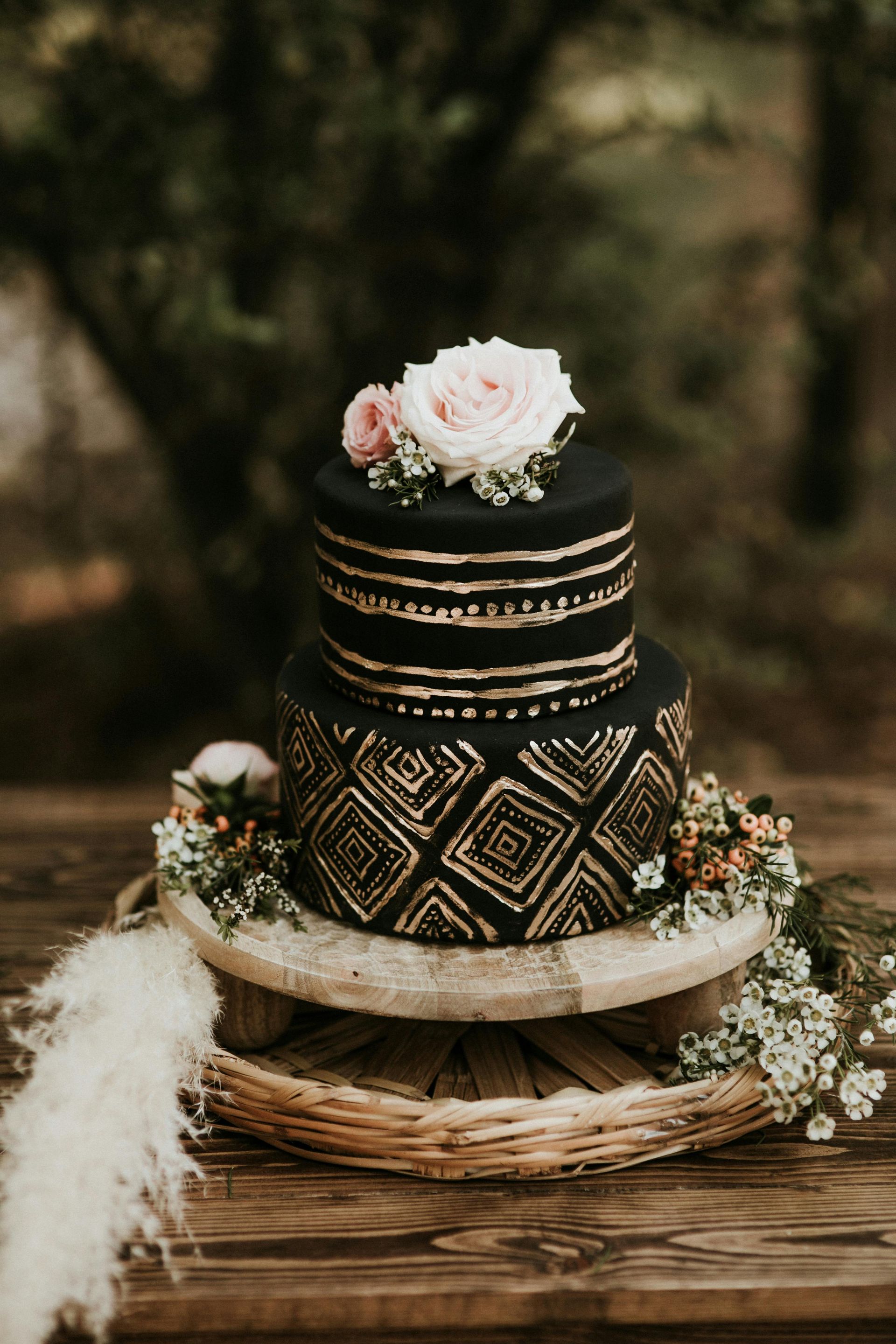 Two-tiered black and gold cake with floral decor on a wooden stand in a natural setting.