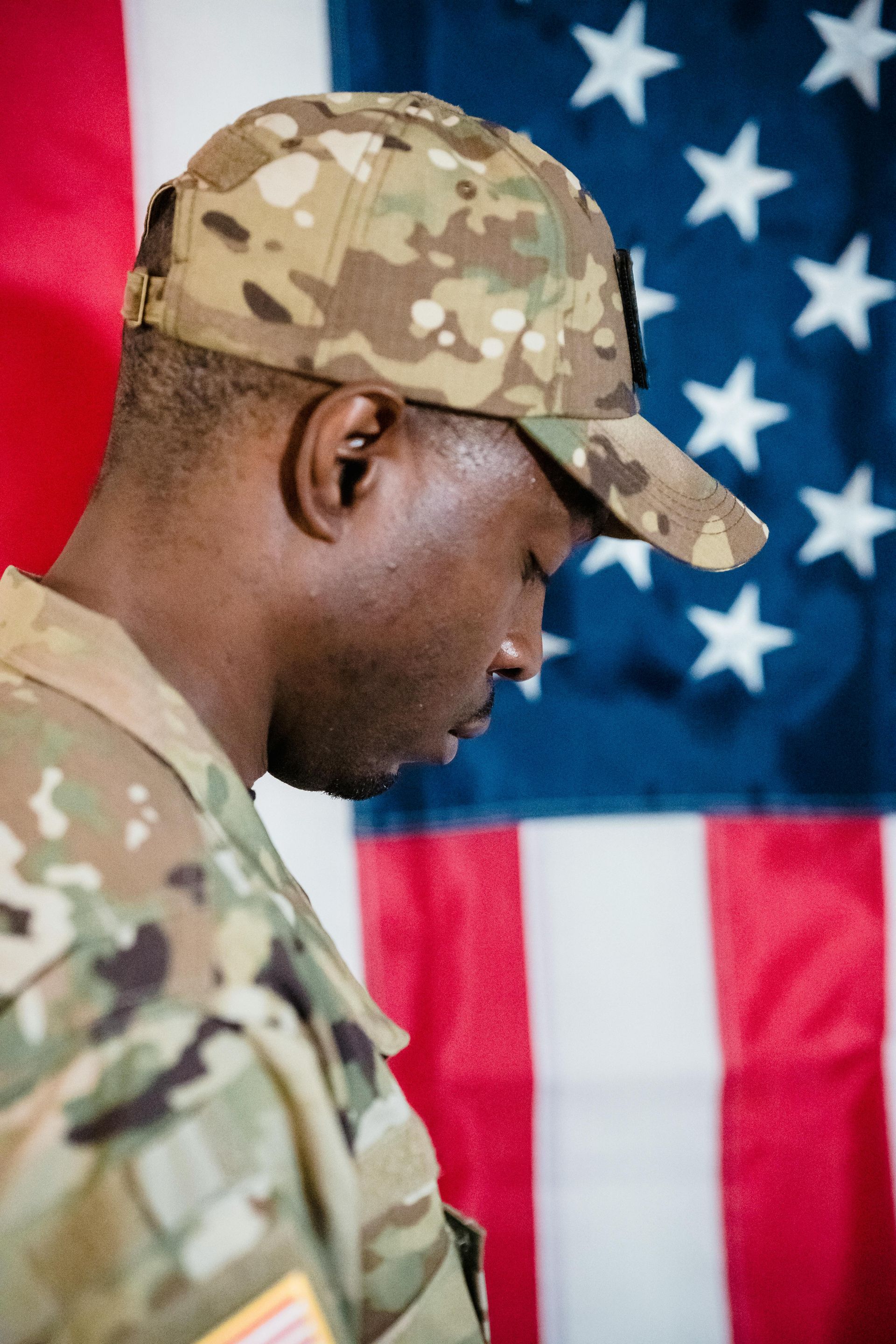 Soldier in camouflage cap and uniform looks down in front of an American flag.