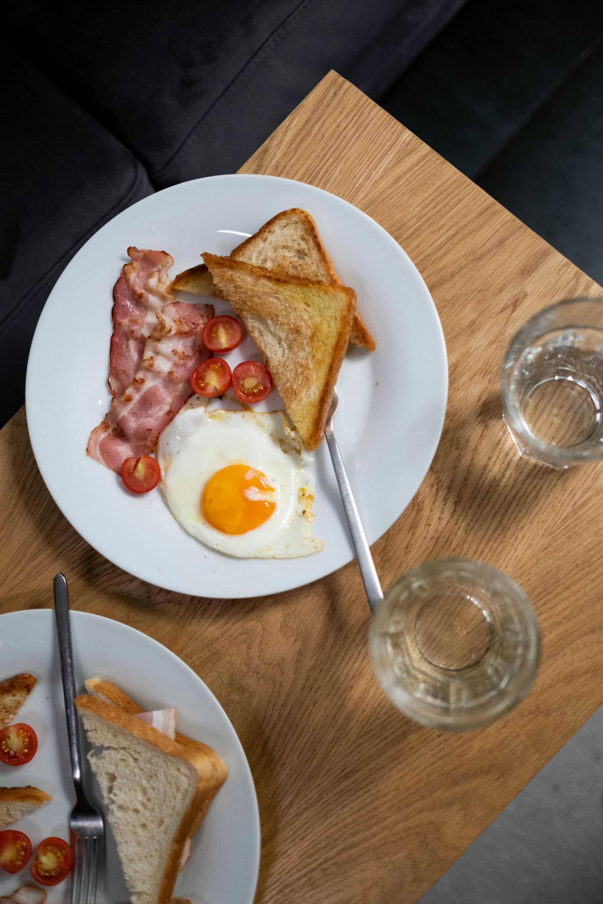 Breakfast plate with bacon, egg, tomatoes, toast; adjacent plate with toast and tomatoes, two glasses of water.