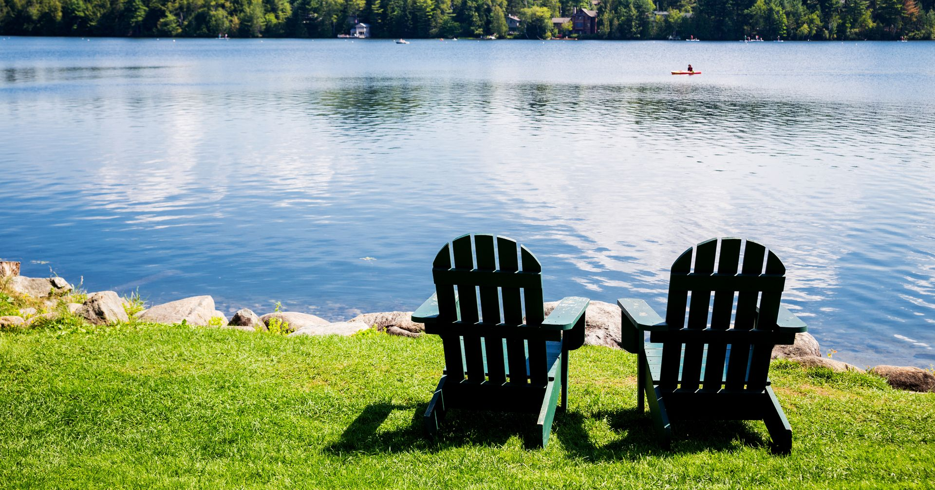 Two dark green chairs on a grassy lakeshore facing calm water and a tree-lined opposite shore.