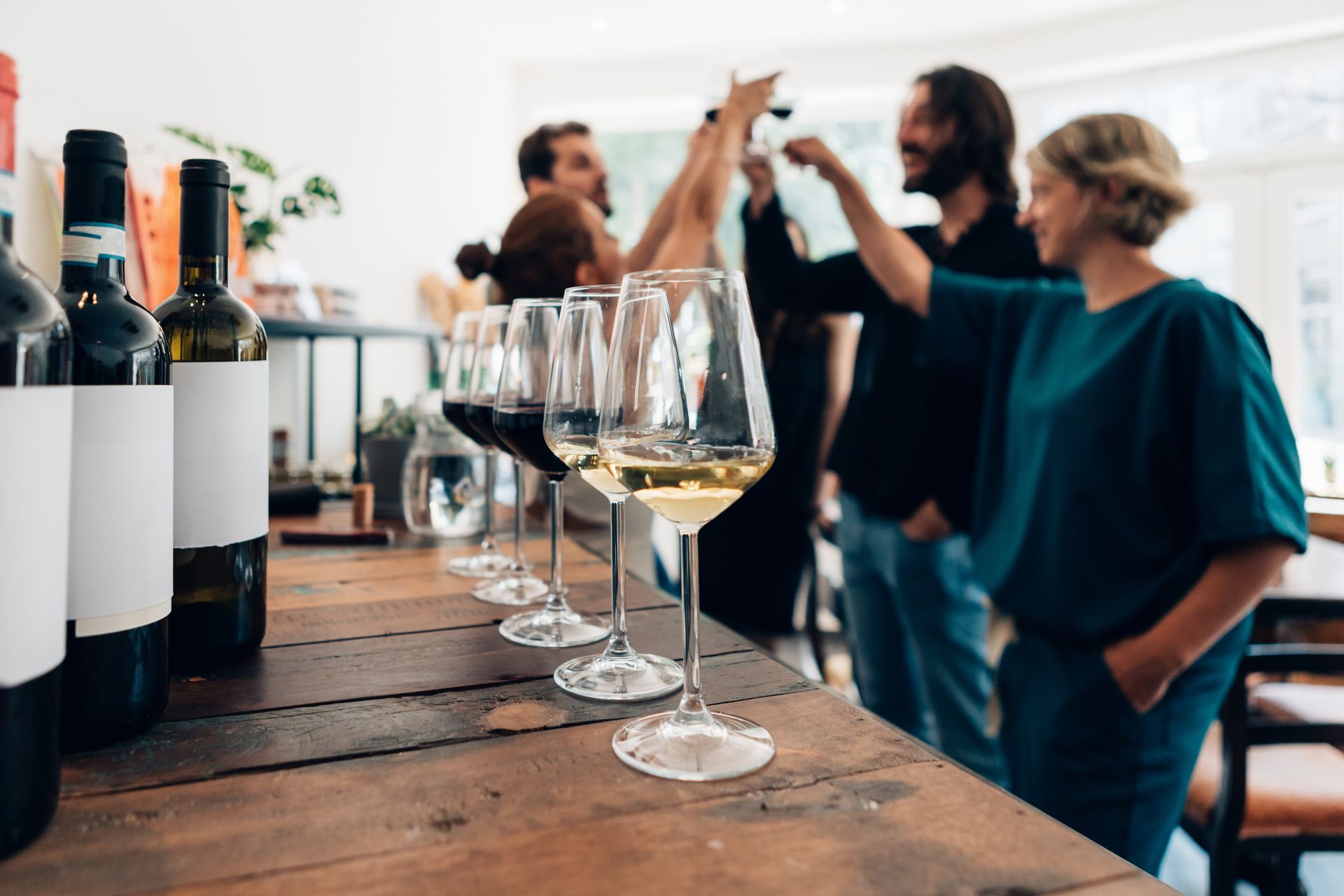 A group of people cheers with wine glasses at a rustic wooden table featuring several wine bottles.