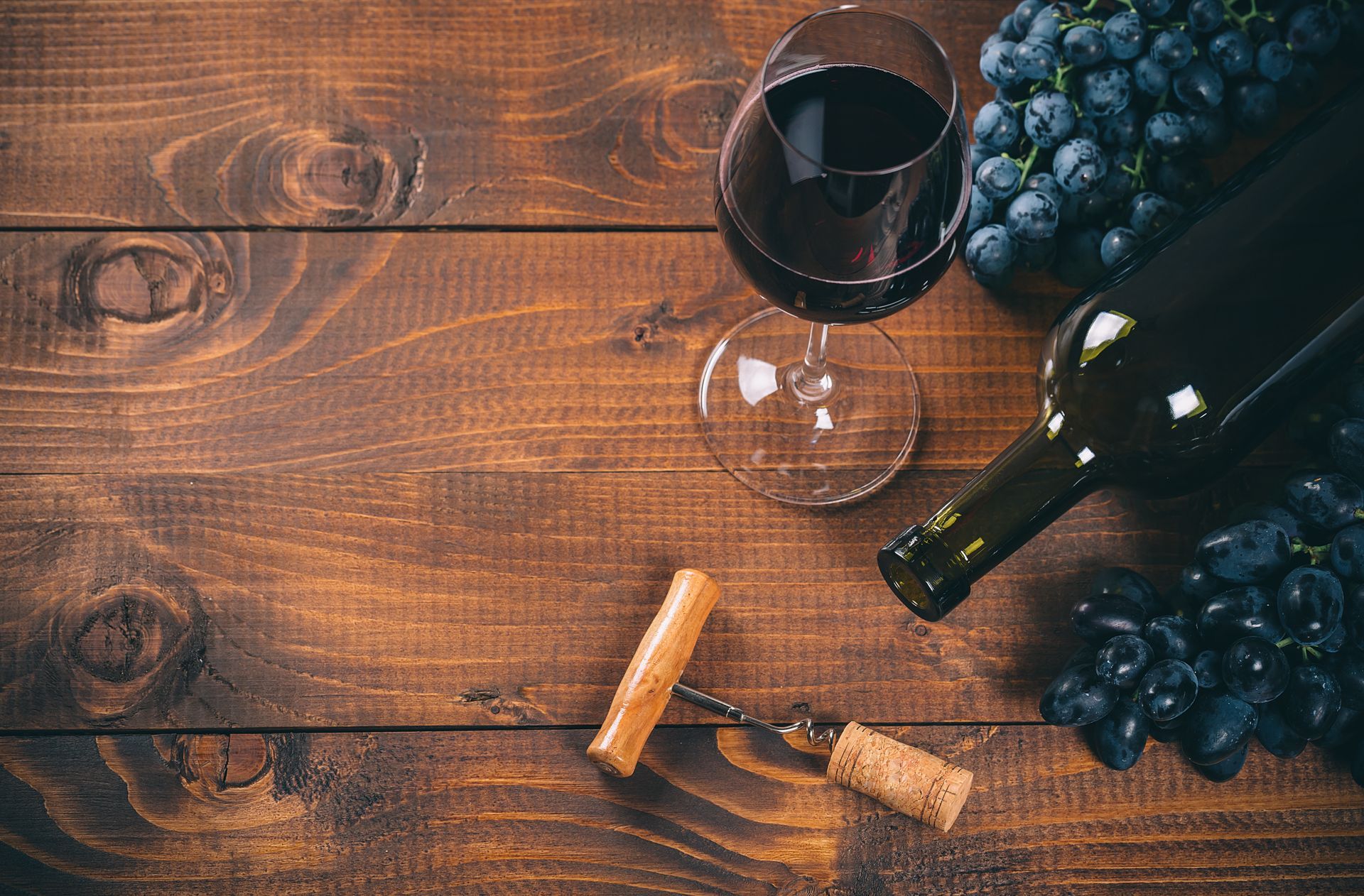 A glass of red wine, a wine bottle, and bunches of grapes on a dark wooden surface with a corkscrew.