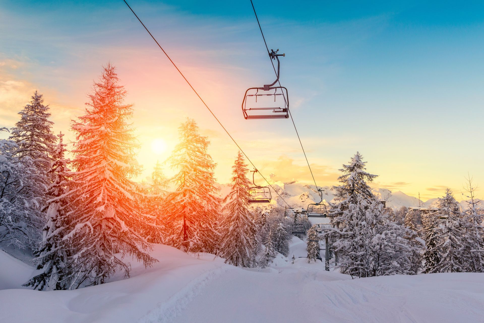 Snowy forest at sunset with ski lift chairs overhead and a glowing sun in a pink-orange sky