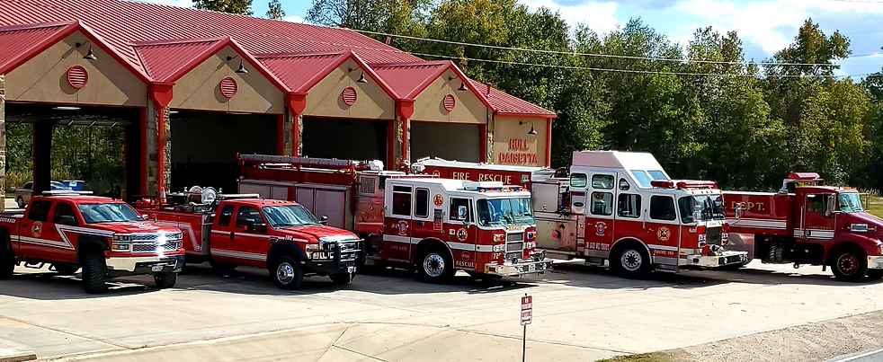 Fire station with red fire trucks parked in front of open garage doors. Trees in background.