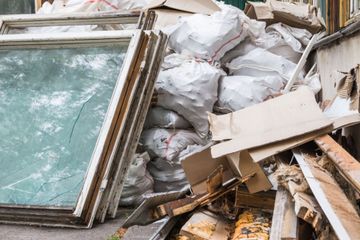 A pile of construction debris, including old window frames, stacked white waste bags, and discarded cardboard and wood.