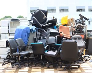 A large, cluttered pile of various office chairs, including black, brown, and yellow models, sits outdoors on a paved area.