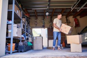 A person standing barefoot in a garage, placing a cardboard box onto a stack of boxes. Shelving units are nearby.