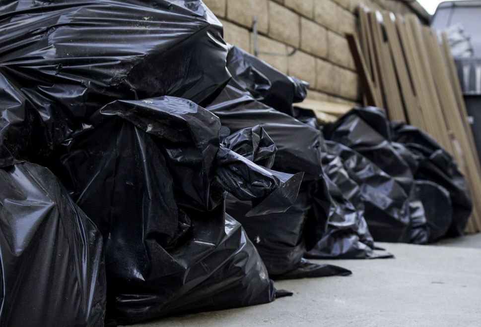 A pile of black garbage bags stacked against a brick wall and wooden planks on a paved ground.