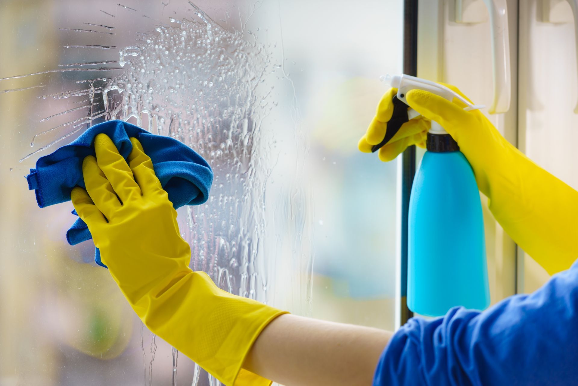 A person wearing yellow gloves is cleaning a window with a cloth and spray bottle.